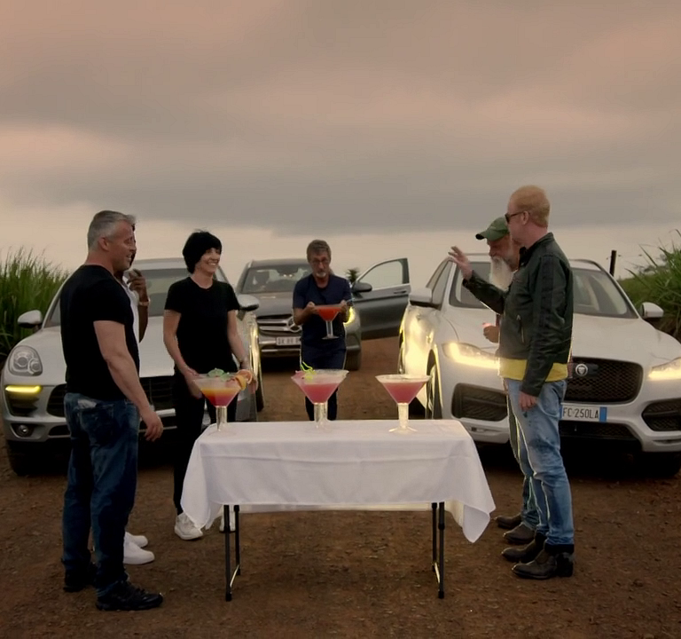 Five people standing outdoors around a table with large colorful cocktails, cars parked behind them, and an overcast sky.