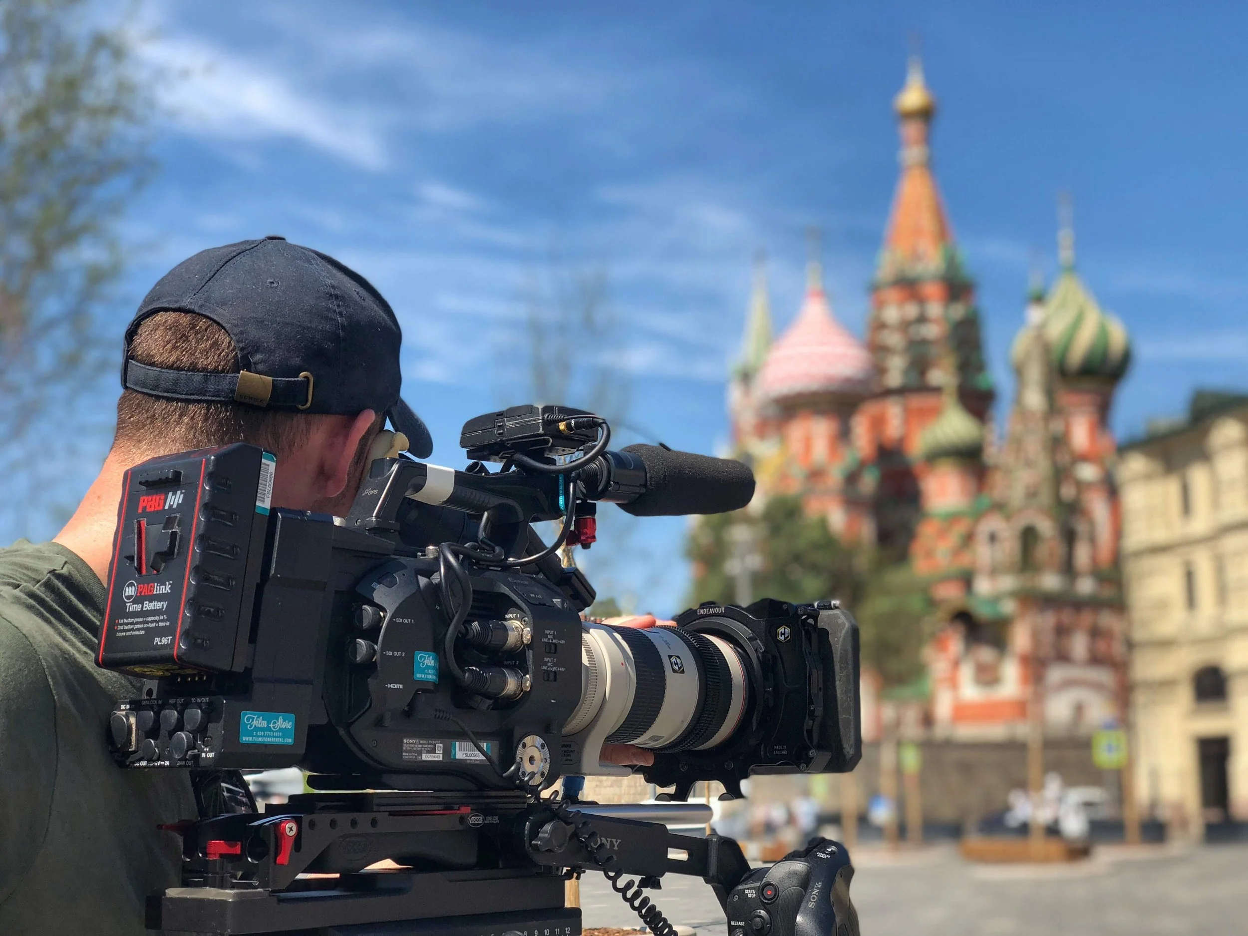 A person operating a professional video camera outdoors with a castle in the background. The person is wearing a black cap, and the camera is mounted on a rig.
