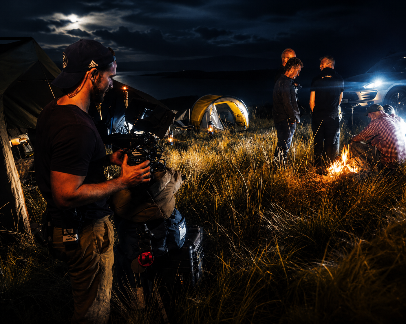 Group of people camping outdoors at night, some around a small campfire, with tents and a car nearby, under a dark sky with clouds.