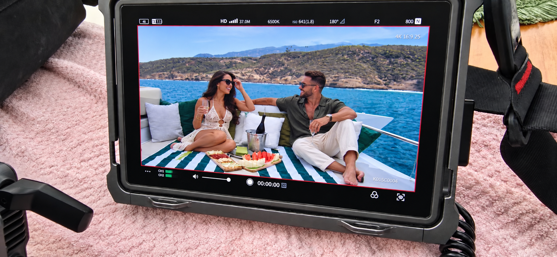 A camera monitor shows a photo of a man and woman relaxing on a boat with scenic water and hills in the background, enjoying drinks and snacks.