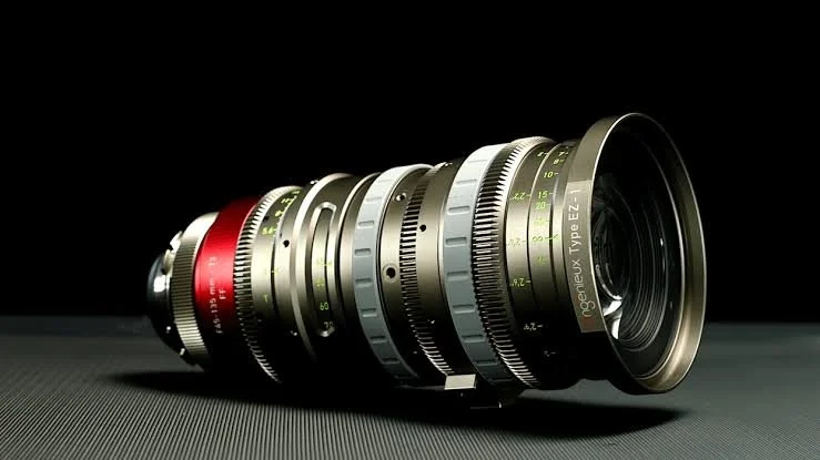 Close-up of a professional camera lens with multiple rings and dials, positioned on a dark surface against a black background.