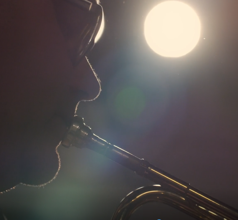 Silhouette of a person playing a wind instrument, possibly a trumpet, with a bright sun or moon in the background.