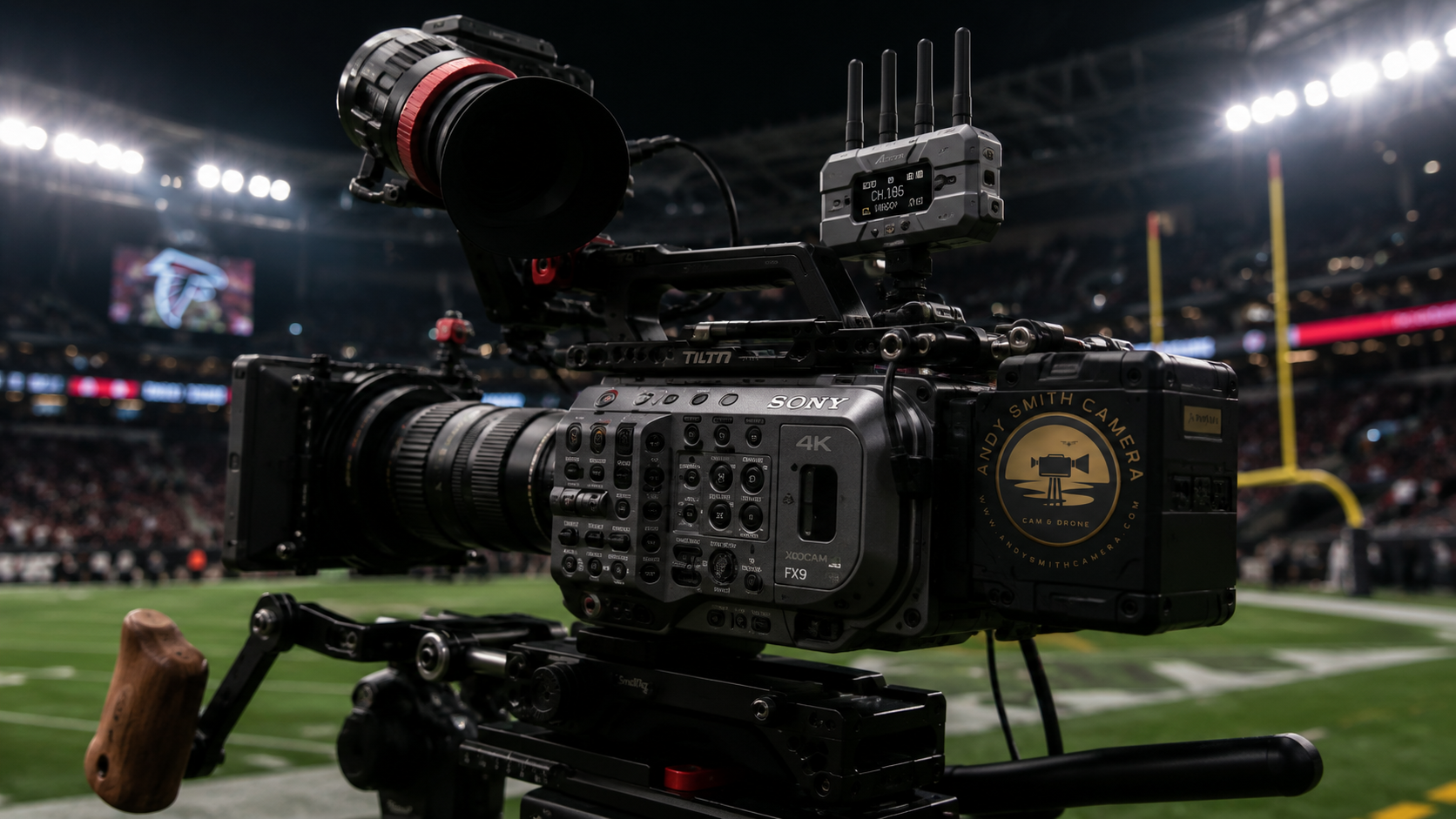 Professional video camera with a large lens and external monitor, positioned on a football field with stadium lights and scoreboard in the background.