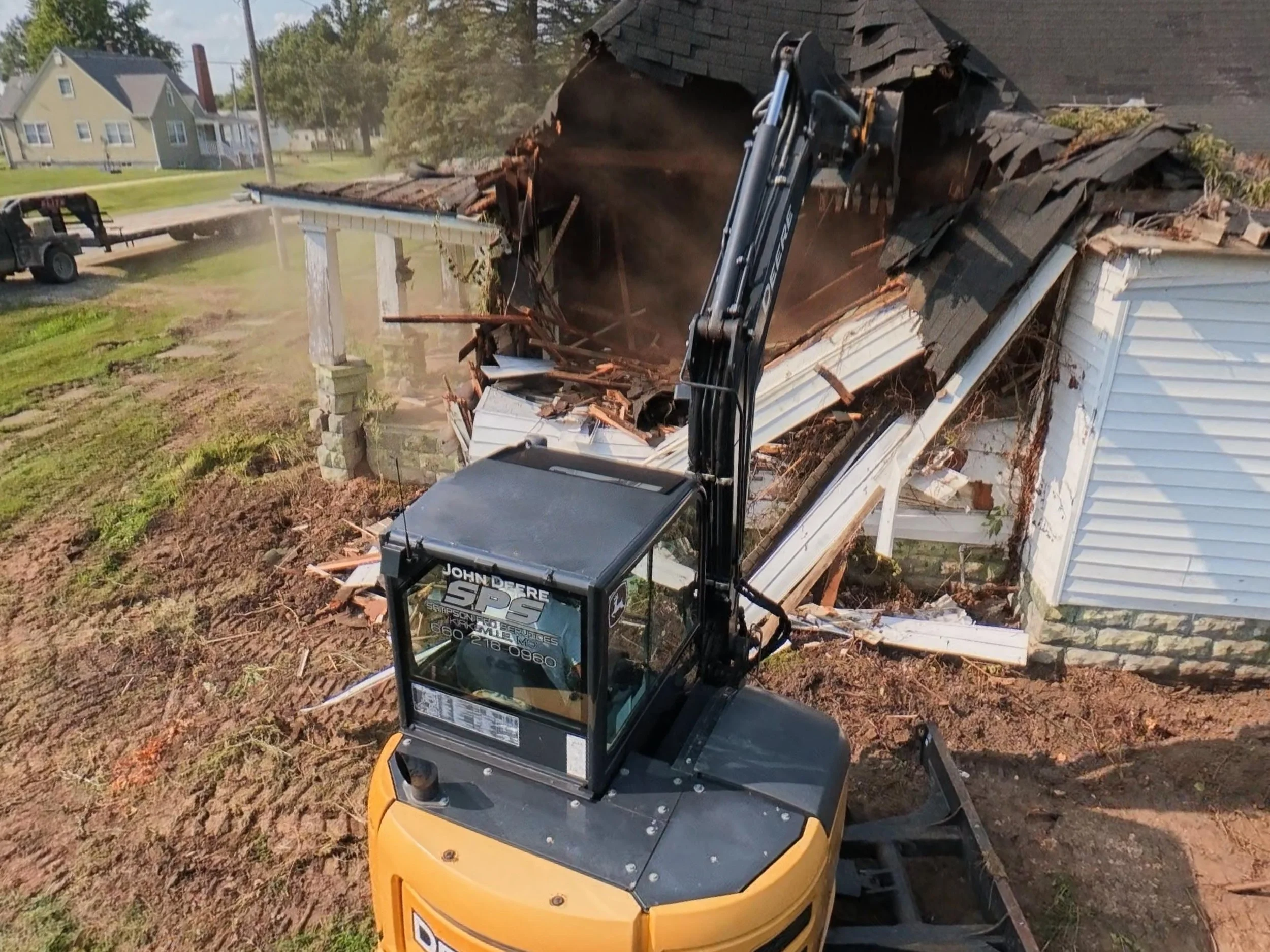 A construction excavator demolishing a small house or garage, with debris falling as the structure is torn down.
