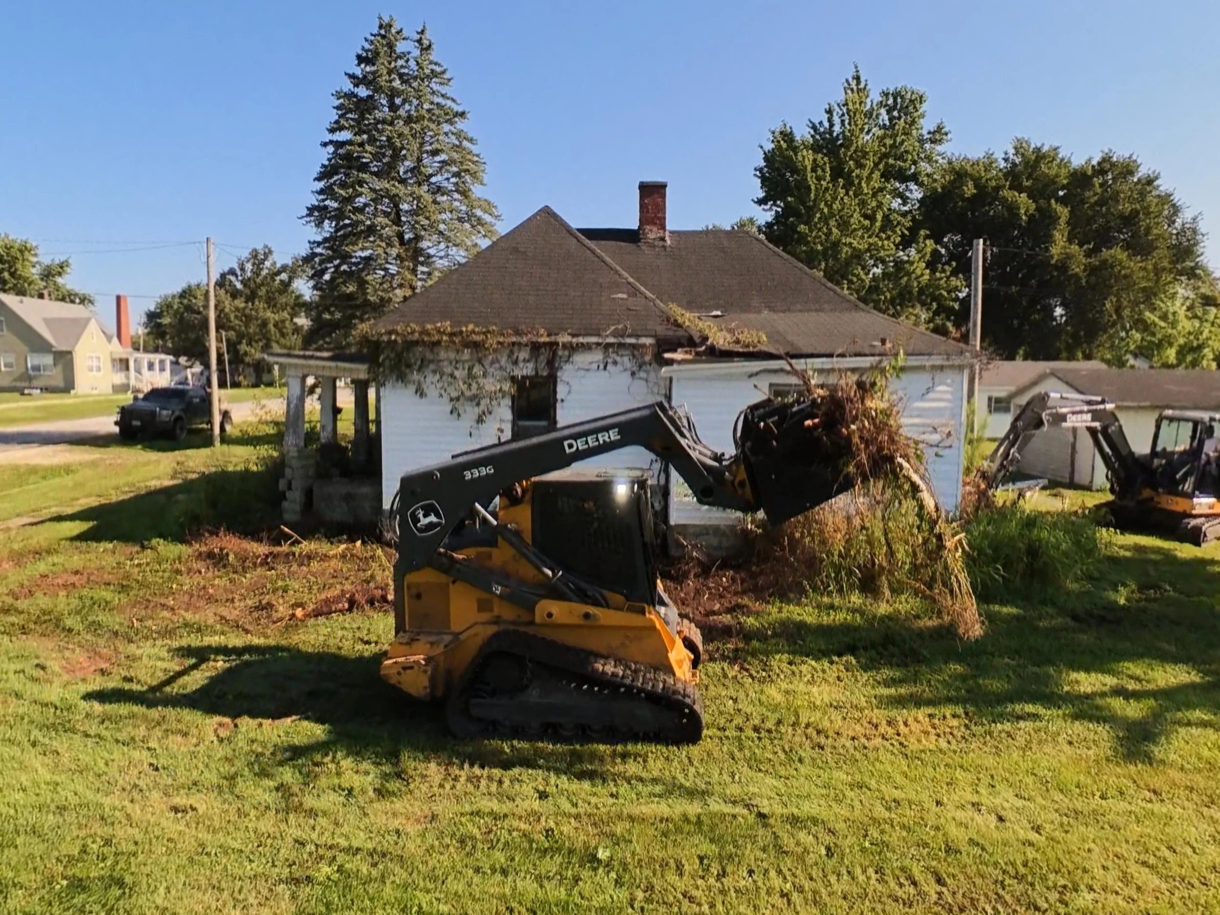 A white house with a dark roof showing signs of damage and overgrown vines, with a yellow mini excavator in the foreground and a black pickup truck in the background, surrounded by green grass and trees under a clear blue sky.