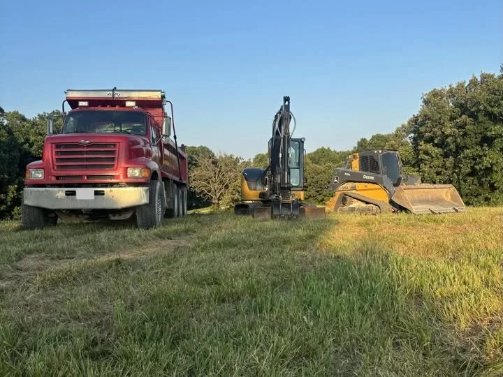 A red dump truck and two construction vehicles, an excavator and a bulldozer, parked on a grassy field with trees in the background during daytime.