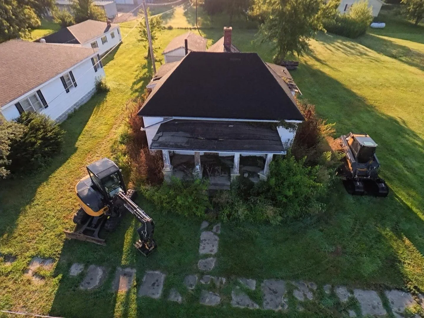Aerial view of an old house undergoing renovation, with construction equipment like a small excavator and a bulldozer nearby, surrounded by green grass and trees.