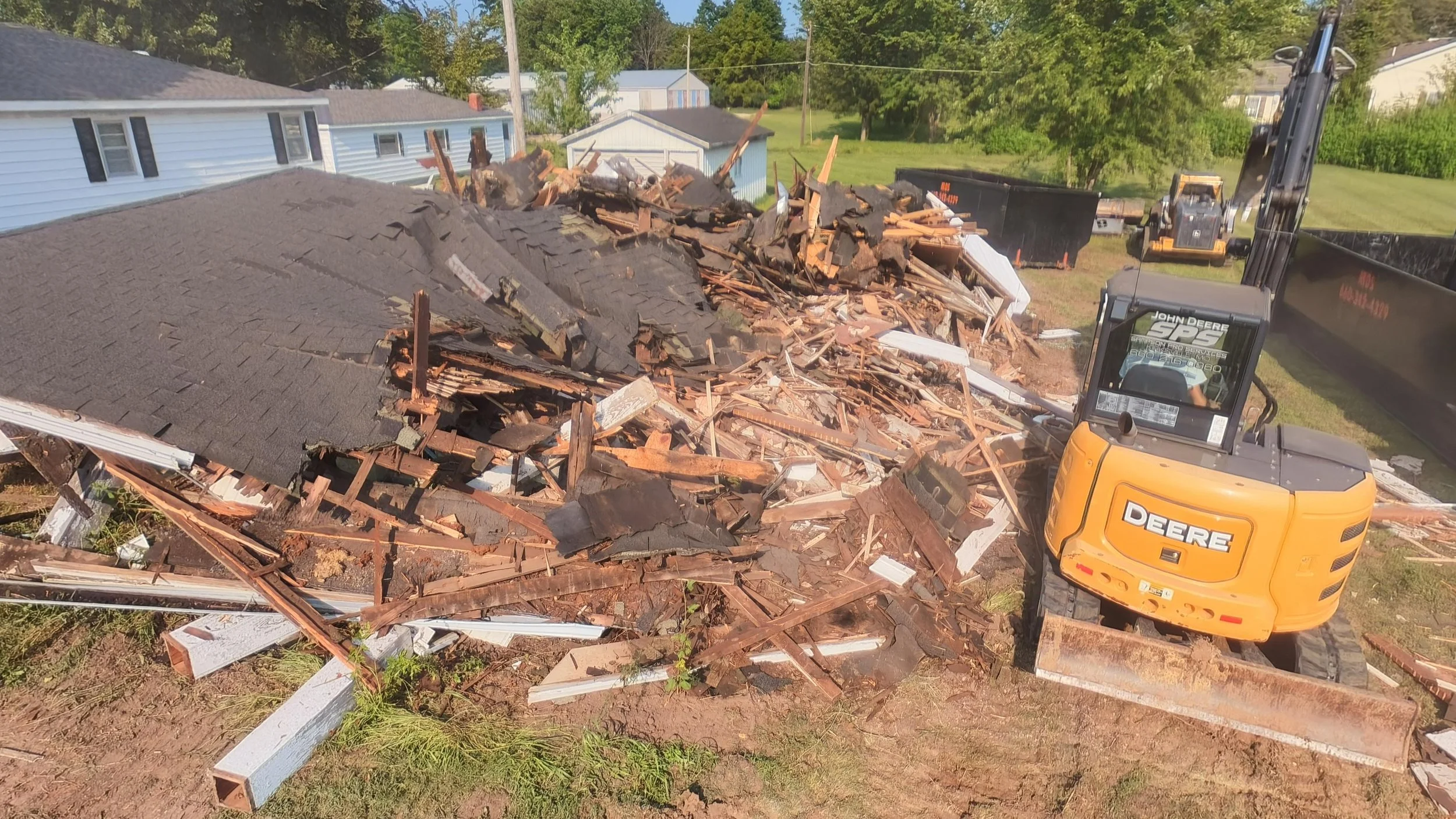An aerial view of a house roof toppled by a demolition, with debris, broken wood, and roofing materials scattered. A yellow John Deere excavator is working on the site. There are neighboring houses and green trees in the background.