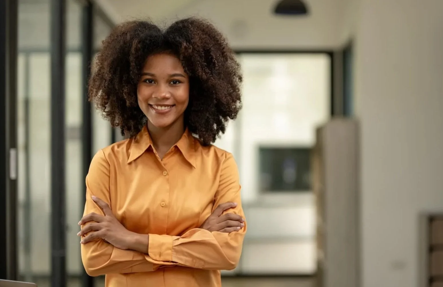 A woman with curly hair wearing an orange button-up shirt, standing with arms crossed and smiling in an office setting.