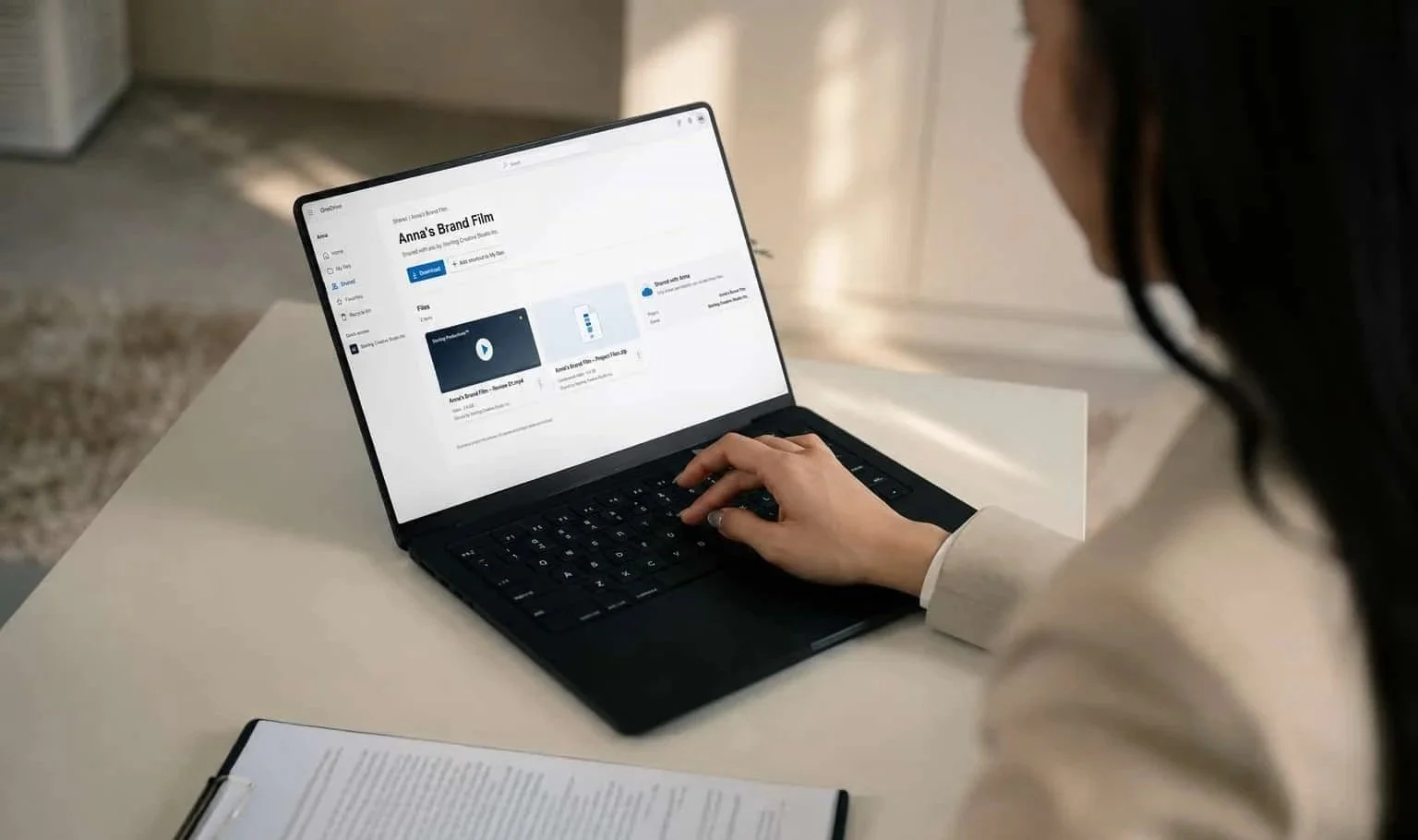 A woman sitting at a desk using a laptop to download a film titled "Anna's Brand Film" from a cloud storage service.