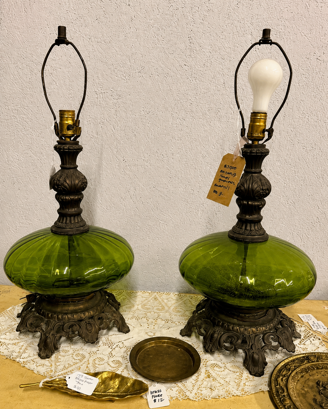 Two vintage table lamps with ornate bronze bases, green glass globes, and exposed light bulb sockets, displayed on a lace-covered table. Sourcing finds from Holly Springs, Raleigh, Sanford, Southern Pines, Pinehurst, Seagrove, High Point, and beyond.