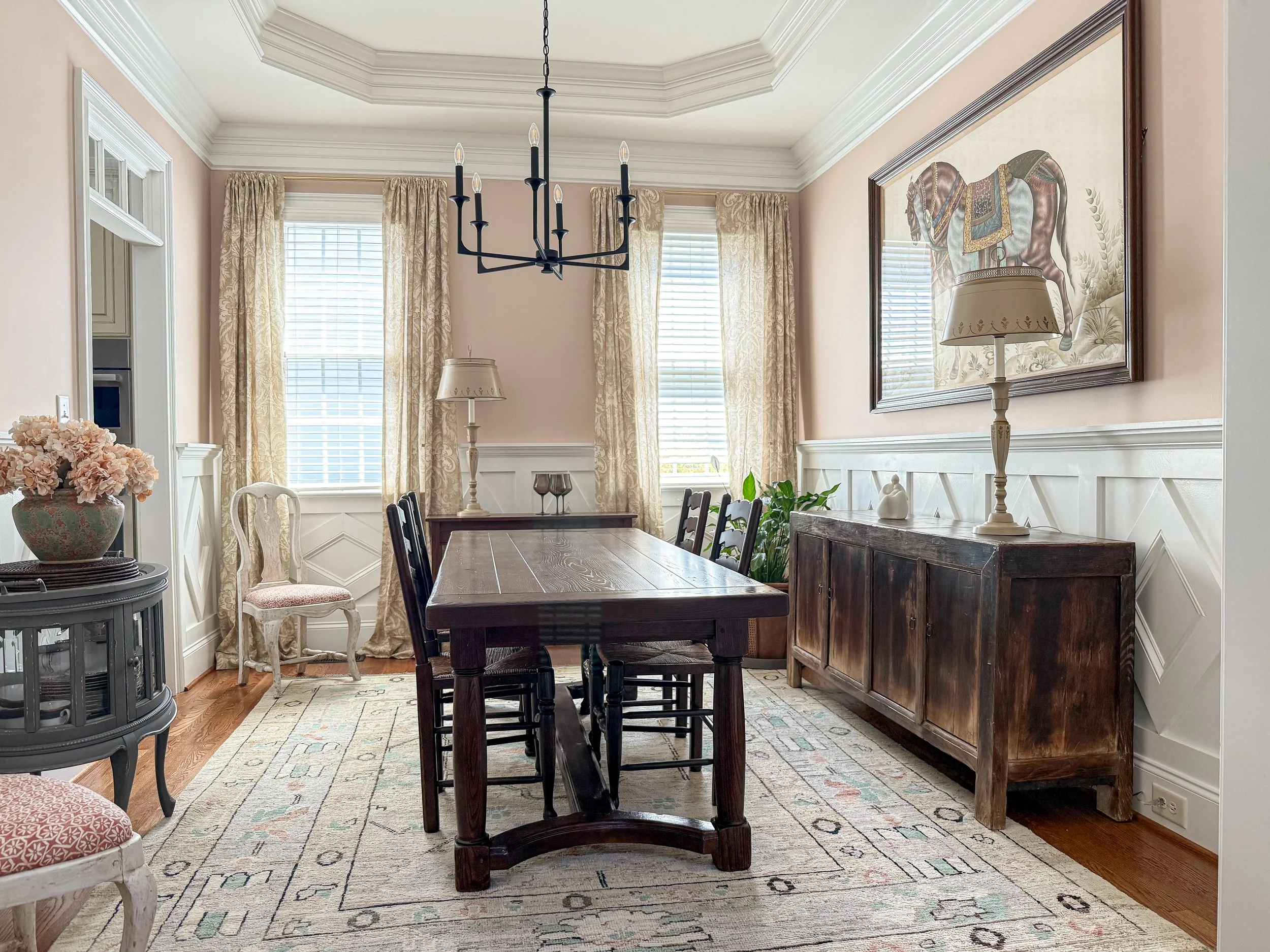 Layered dining room design with an antique wood table, dark chairs, equestrian artwork, chandelier, and styled sideboard.