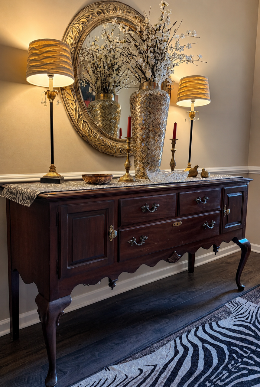 Decorative wooden sideboard with two lamps, a large oval mirror, vases with white flowers, and gold candlesticks with red candles.