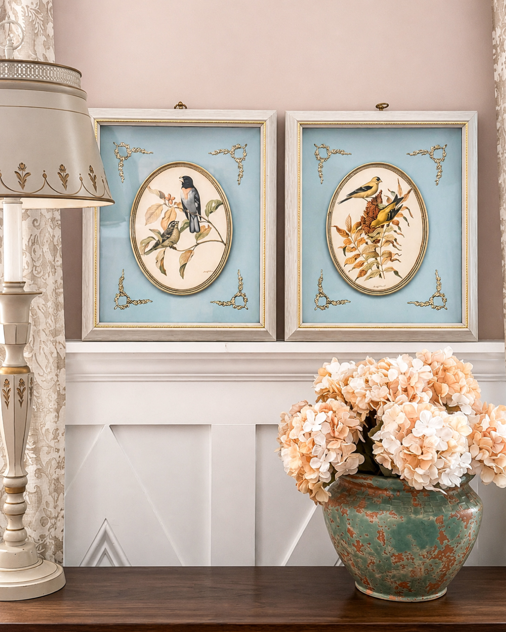 Pair of framed bird prints hanging above a white wainscoting on a pink wall, with a beige floral chandelier on the left and a greenish ceramic vase with peach-colored hydrangeas on a wooden table in the foreground.