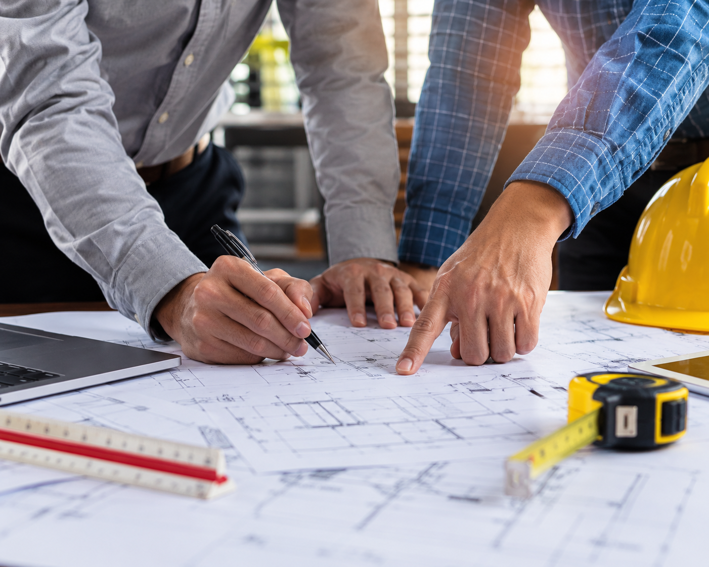 Person in a plaid shirt pointing and drawing on blueprints with a pencil, a white construction helmet and a yellow hard hat on the table.