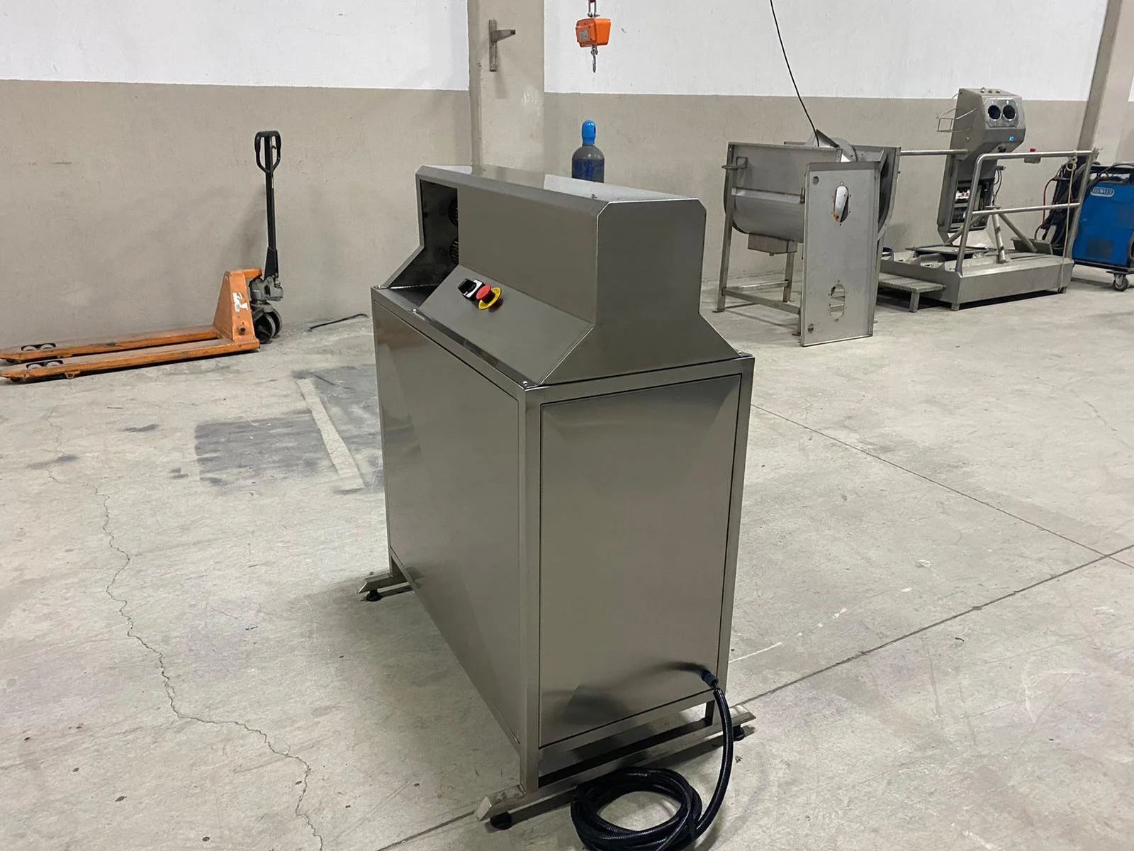 A stainless steel industrial machine with control buttons, in a warehouse or factory with various equipment and tools in the background.