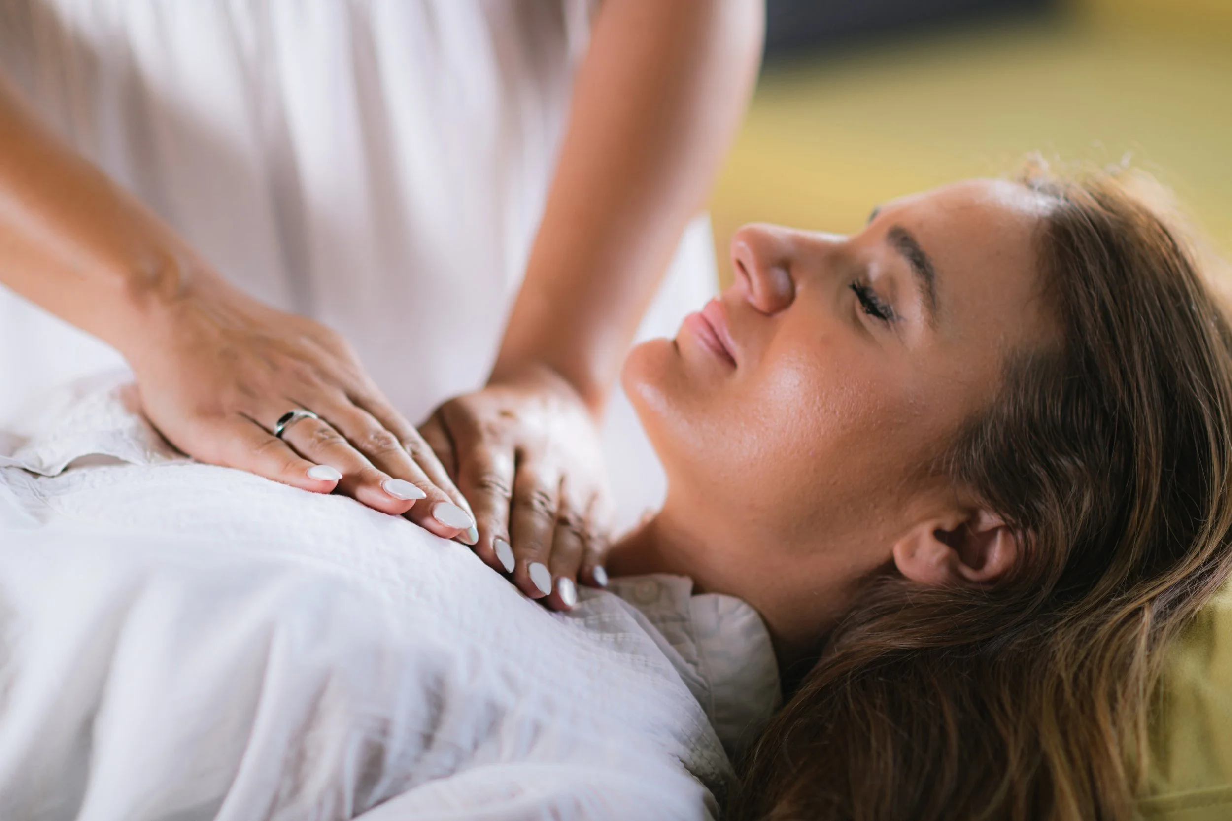 A woman lying on her side with her eyes closed, receiving Reiki on her chest from someone with a blurred natural background.