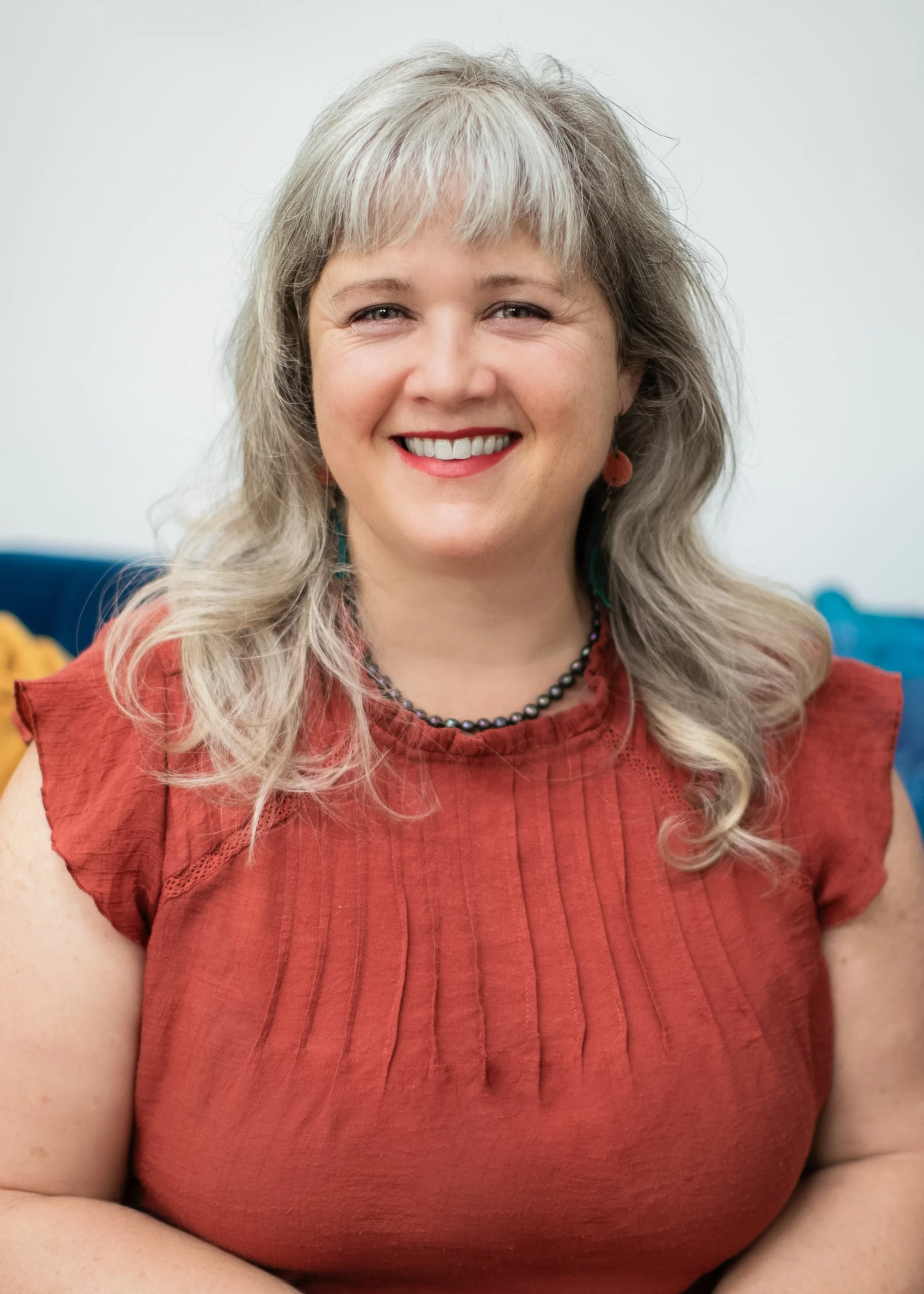 Close-up of a smiling woman with gray hair, wearing a red top and black beaded necklace, sitting in a bright room.