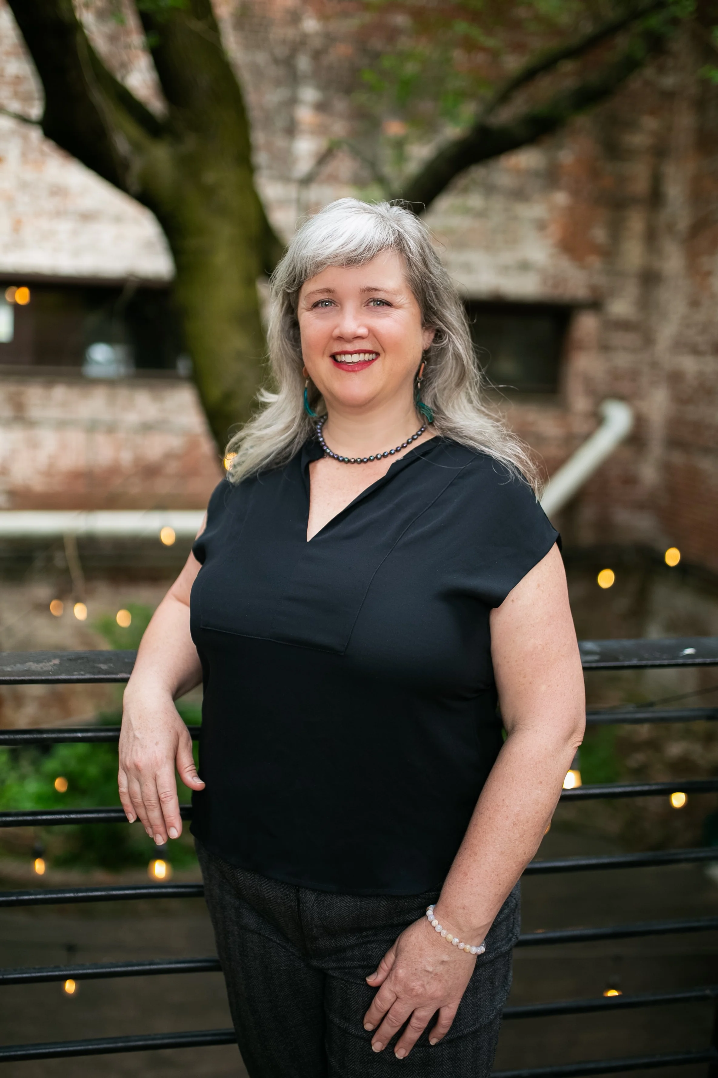 A middle-aged woman with gray hair smiling, wearing a black top, gray checked pants, a black beaded necklace, turquoise earrings, and a pearl bracelet, standing outdoors in front of a brick wall with a large tree and string lights.