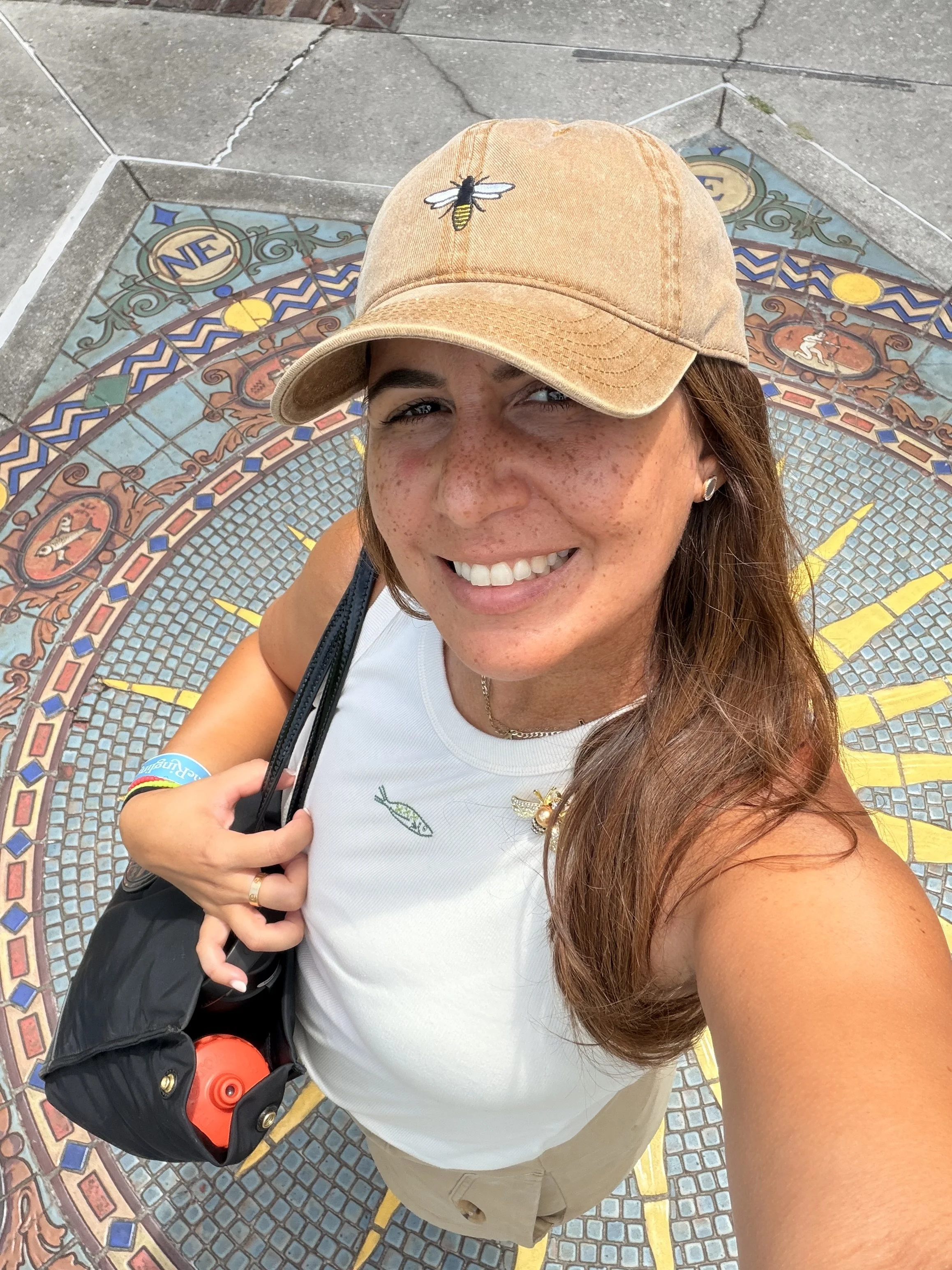 A smiling woman wearing a tan baseball cap with a bee patch, a white tank top, and khaki shorts taking a selfie on a decorative tiled ground with colorful mosaic patterns.