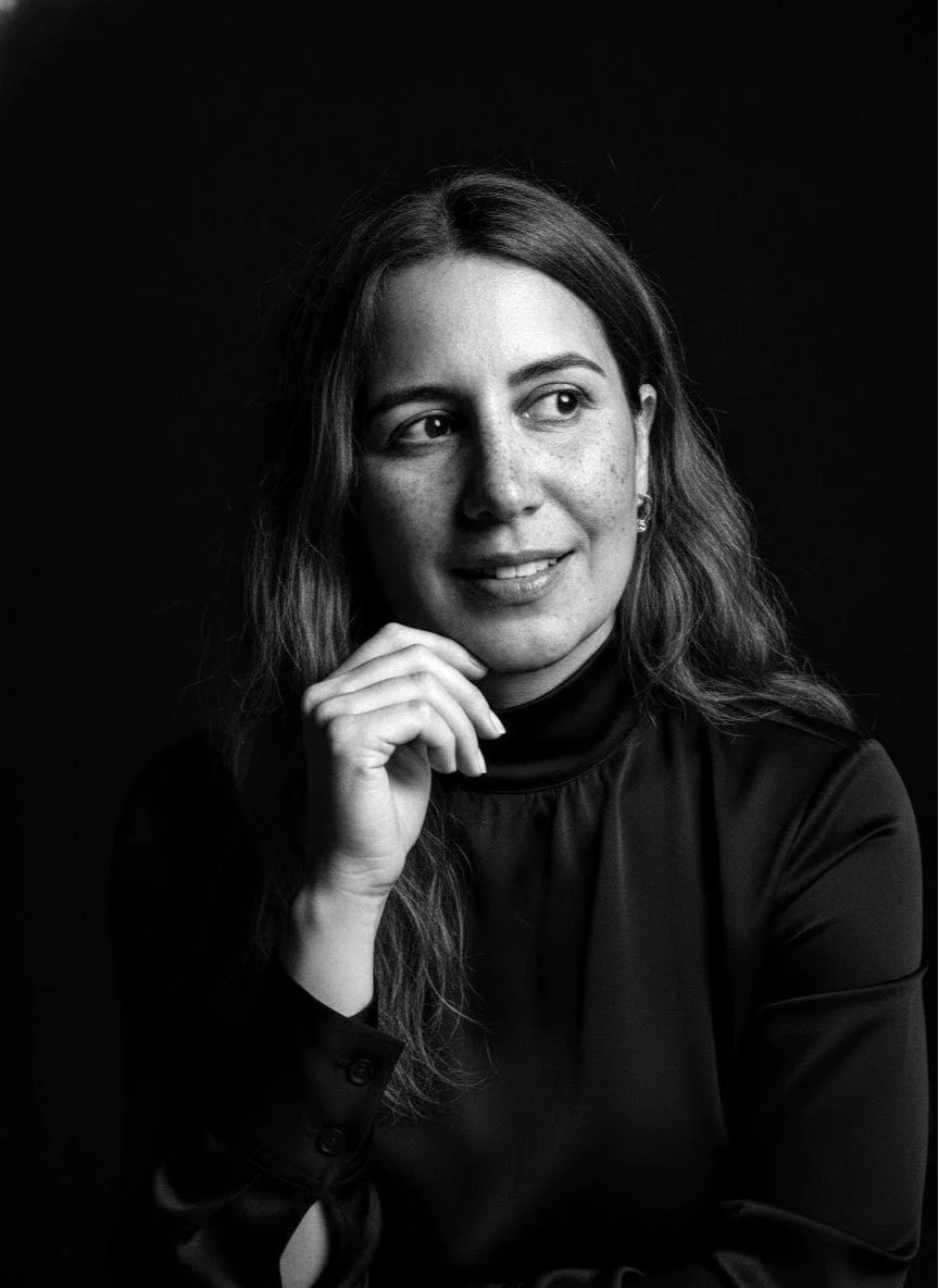Black and white portrait of a woman with long, wavy hair, wearing a turtleneck and earrings, resting her chin on her hand, looking to her left against a dark background.