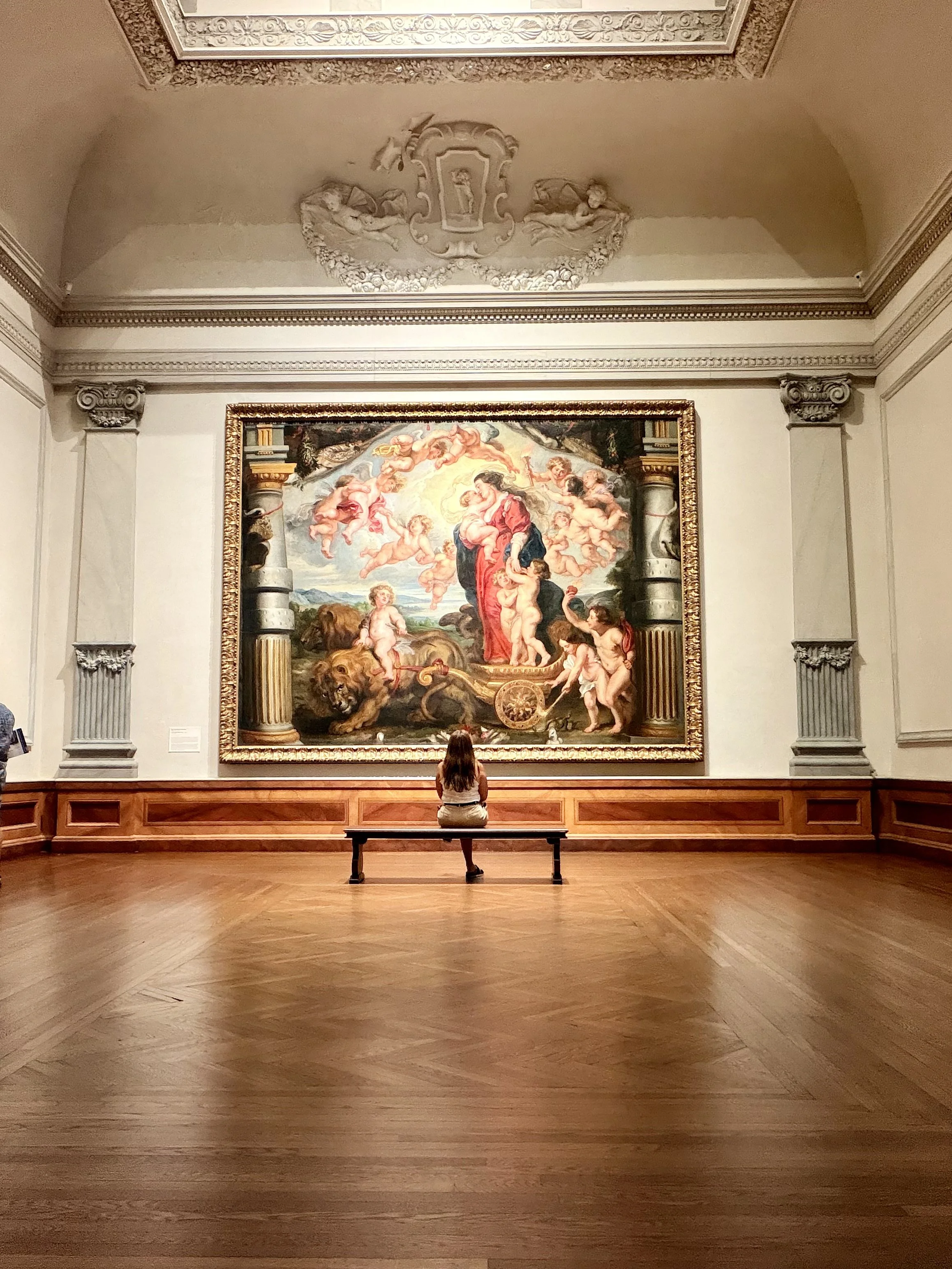 A woman sitting on a bench in an art museum, facing a large framed painting depicting a mythological scene with cherubs, a lion, and a chariot.