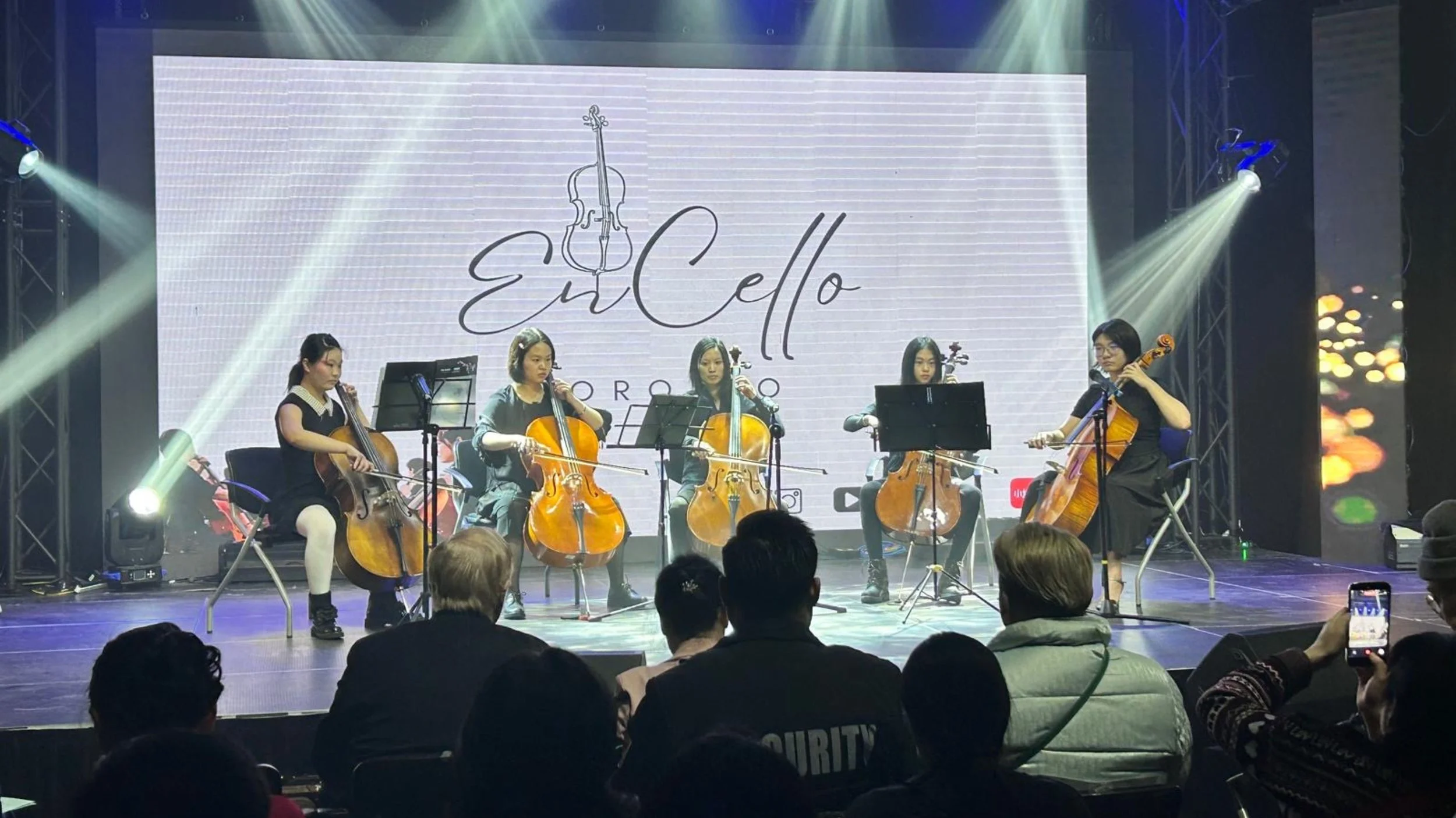A group of five women playing cellos on stage during a performance, with a large screen behind them displaying the words 'Evi Cello' and a drawing of a cello, stage lights, and an audience in the foreground.