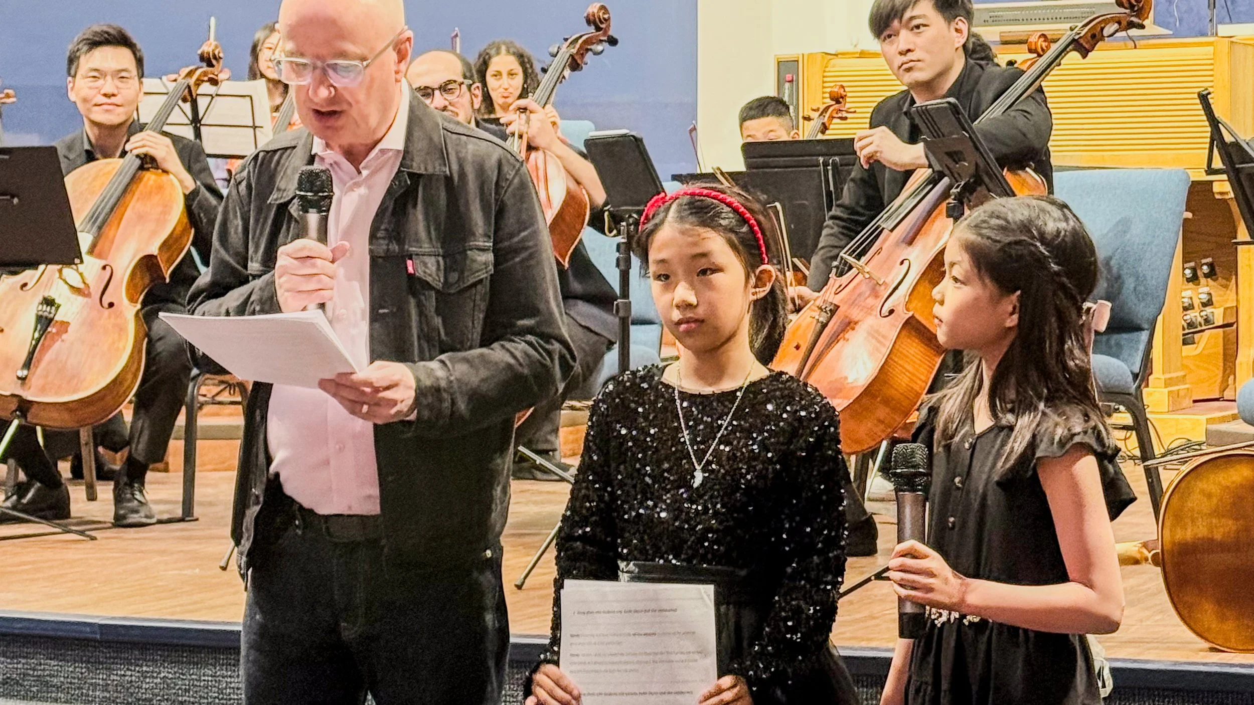 A man reading from a paper while holding a microphone, standing next to two young girls, one holding a microphone, all on a stage with an orchestra of musicians in the background.