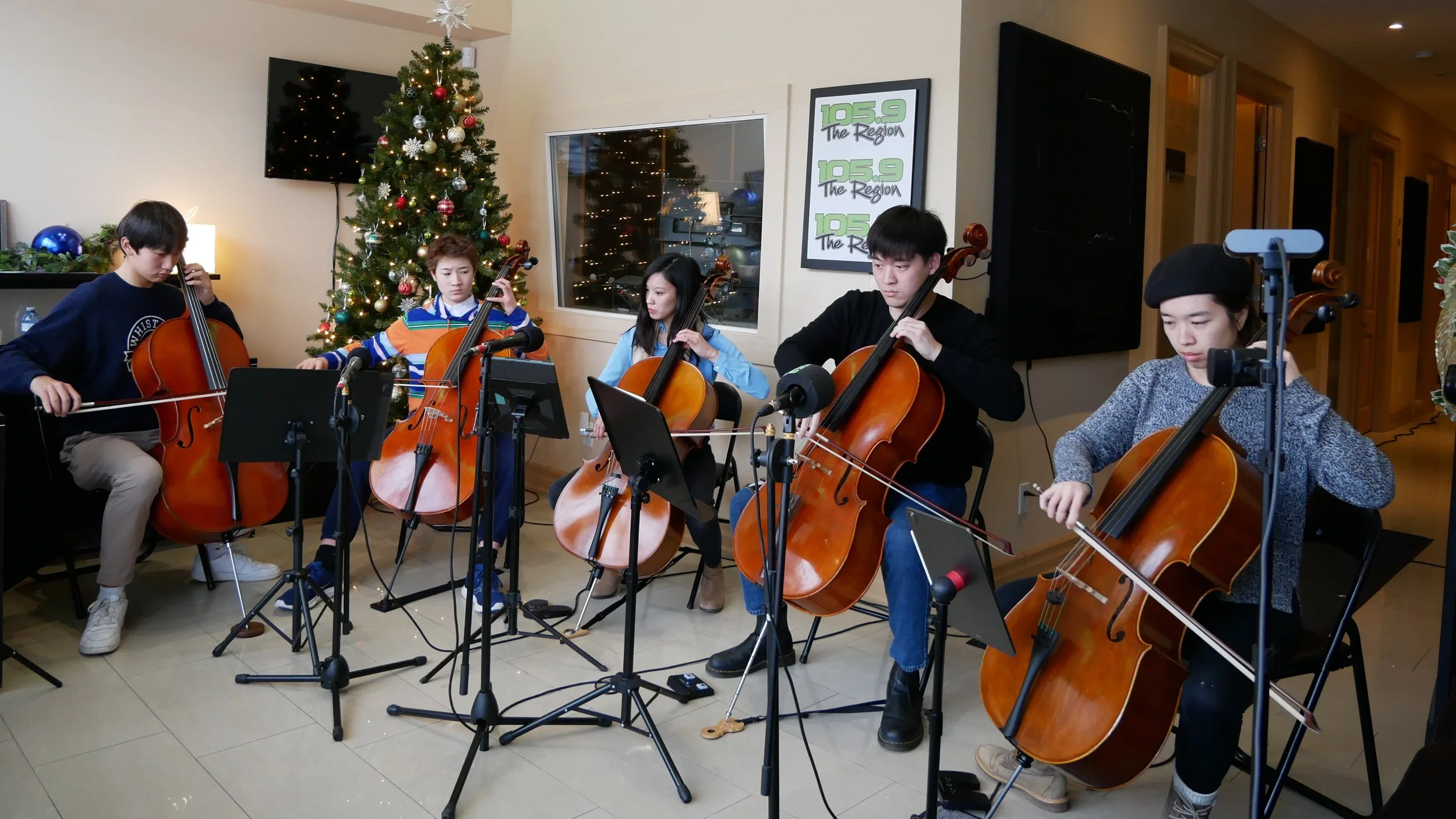 Group of five young musicians playing cellos during a holiday performance in a living room decorated with a Christmas tree and ornaments.
