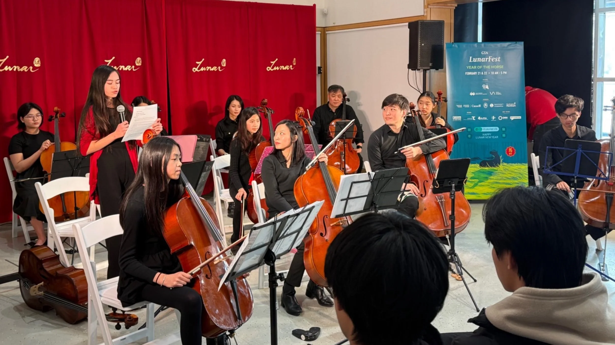 Young woman reads sheet music while holding a microphone during a string orchestra performance, with several musicians playing cellos and violins in the background, red curtain with gold lettering, and a blue poster with event information.