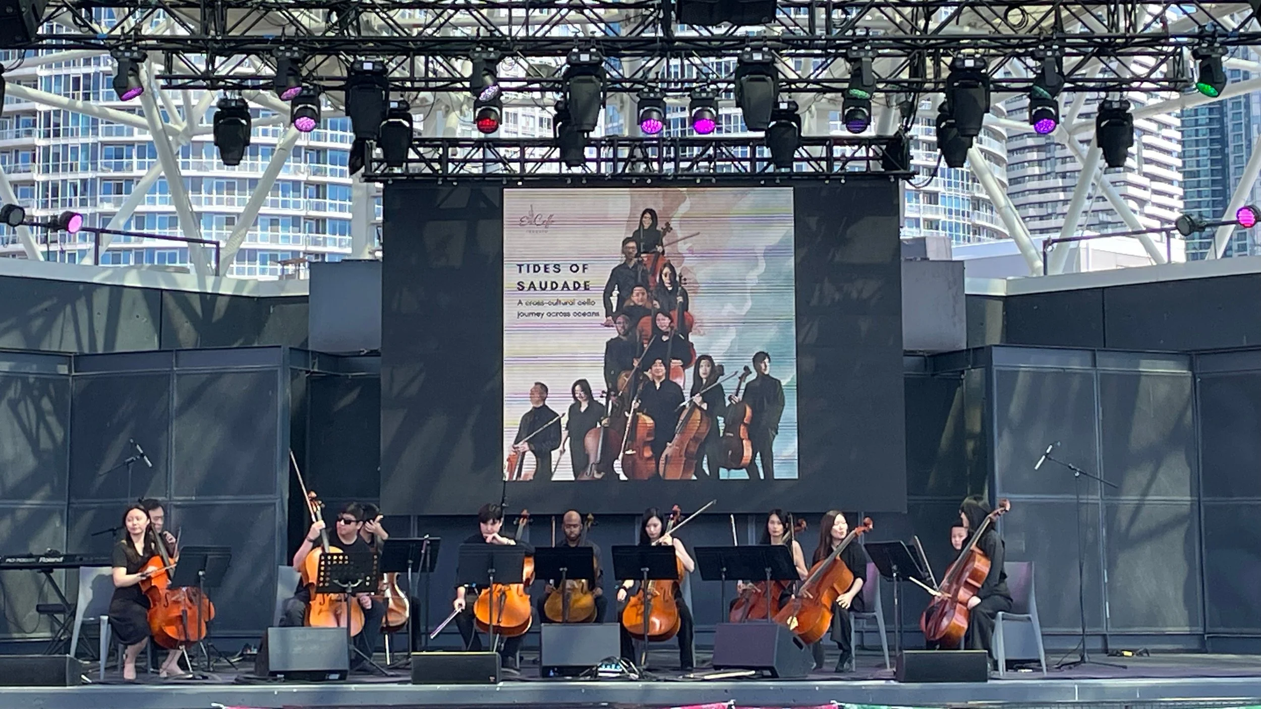 An outdoor concert stage with a large digital screen displaying a promotional poster for 'Tides of Saudade,' a classical music ensemble. The orchestra, composed of string musicians, is performing in front of a cityscape background with tall modern bu