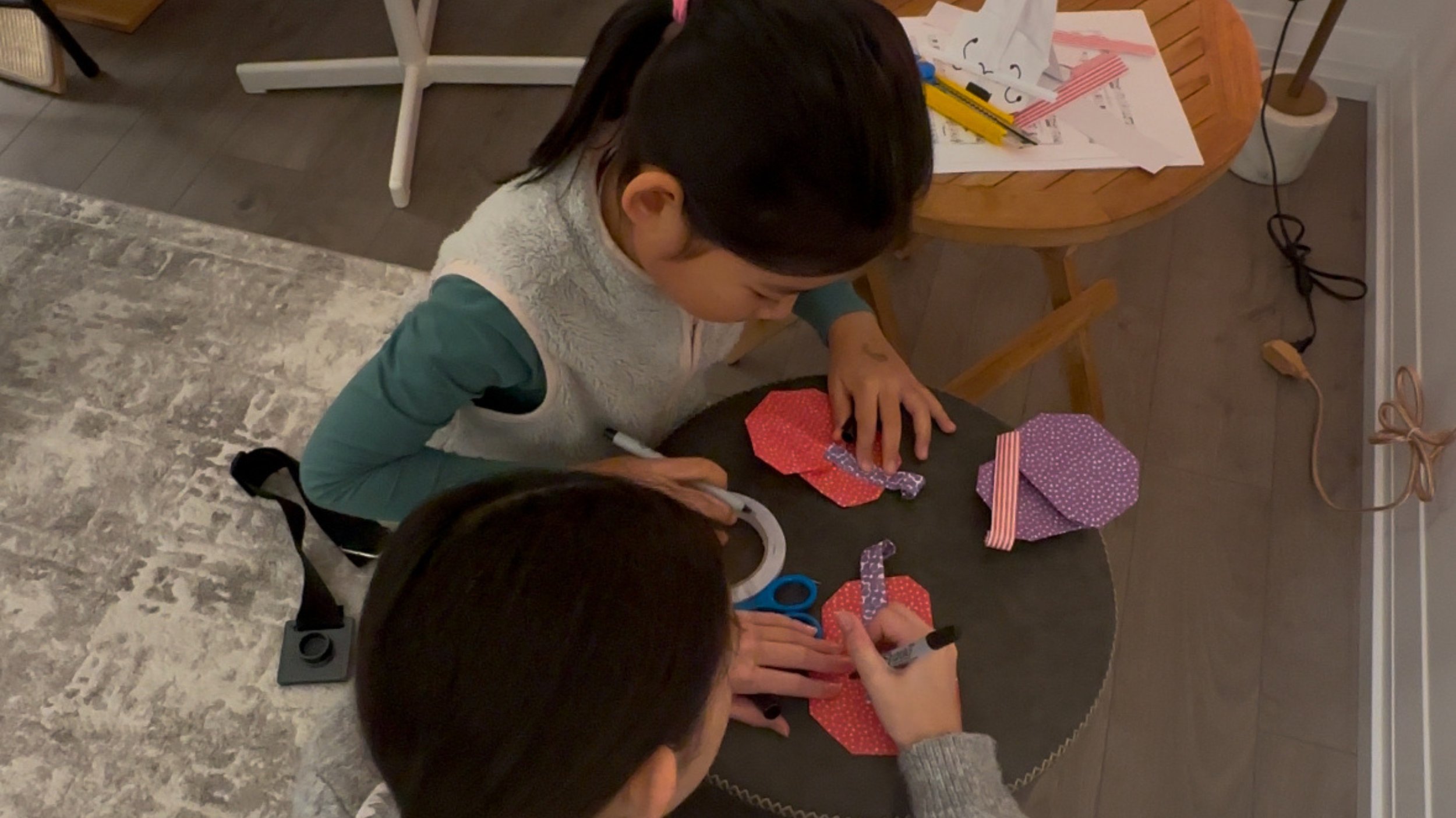 Two children sitting at a table engaging in arts and crafts, cutting and handling colorful paper, in a cozy indoor setting.