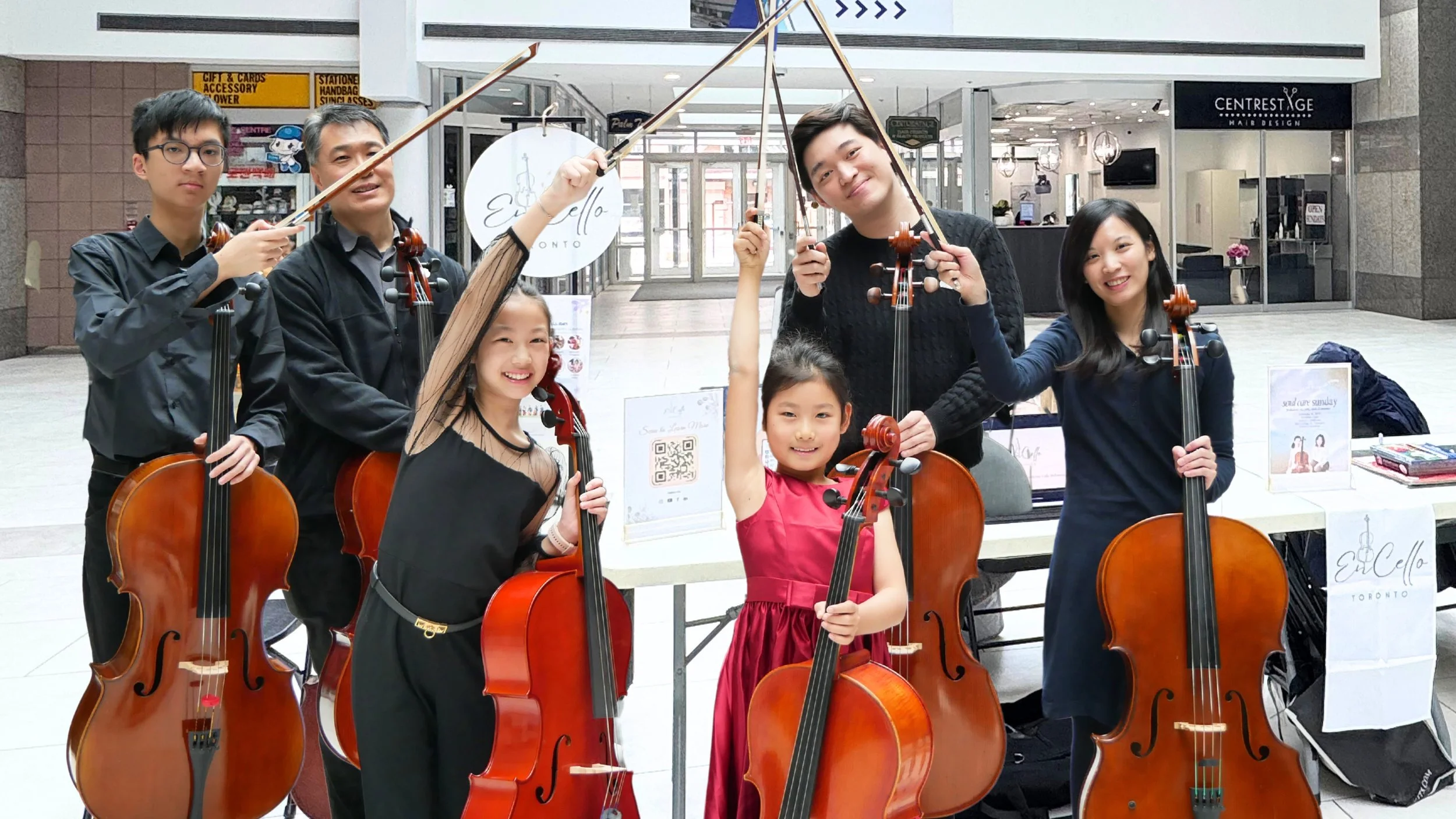 A group of seven people, including children and adults, holding musical cellos and bows, posing together in a shopping mall. They are smiling and standing behind a table with promotional signs.