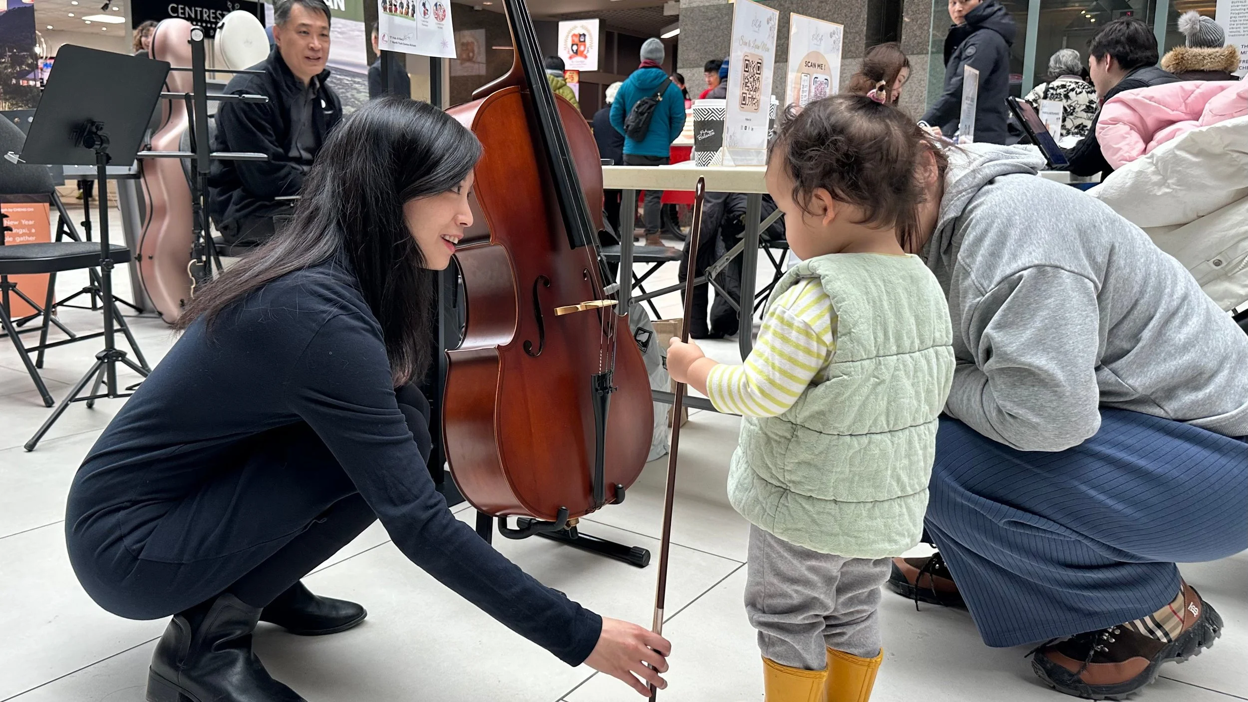 A woman crouching down playing a violin for a young girl at a public event in a shopping mall.