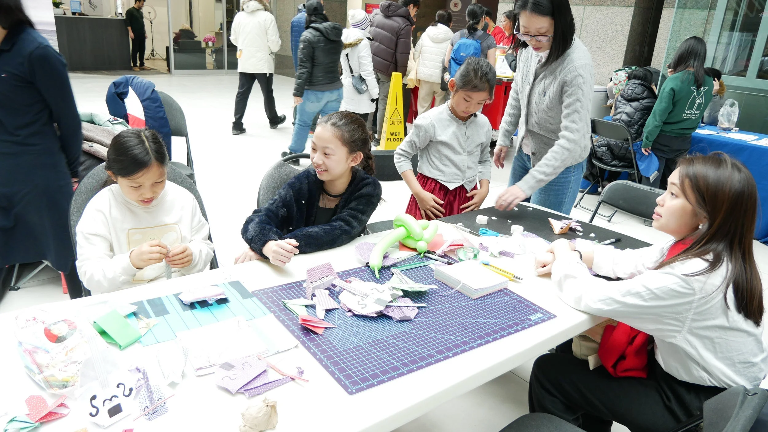 Kids and adults participating in a crafting event at a registration table with various craft supplies, in an indoor public space.