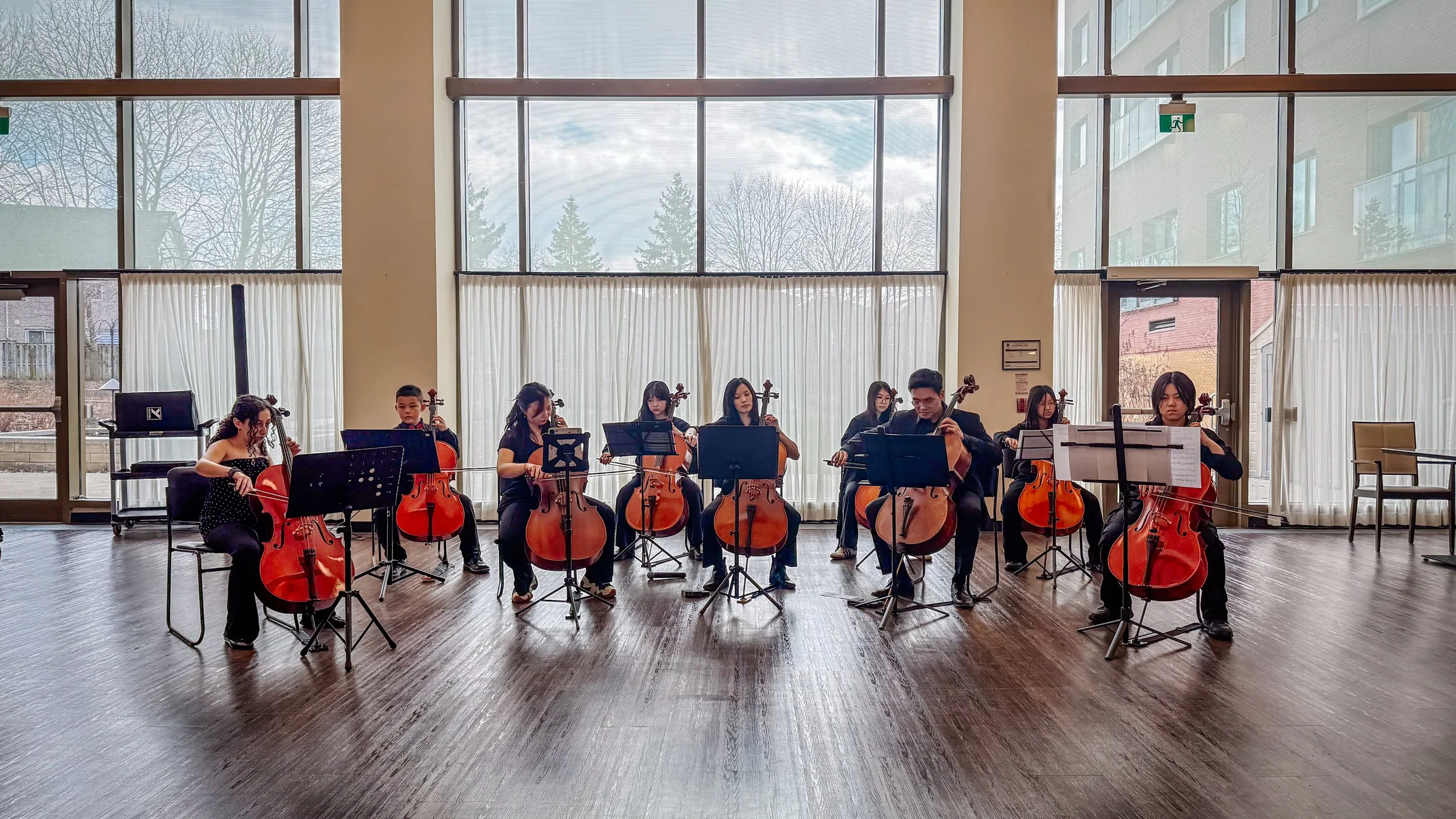 A group of violinists practicing indoors with large windows behind them showing trees and buildings.