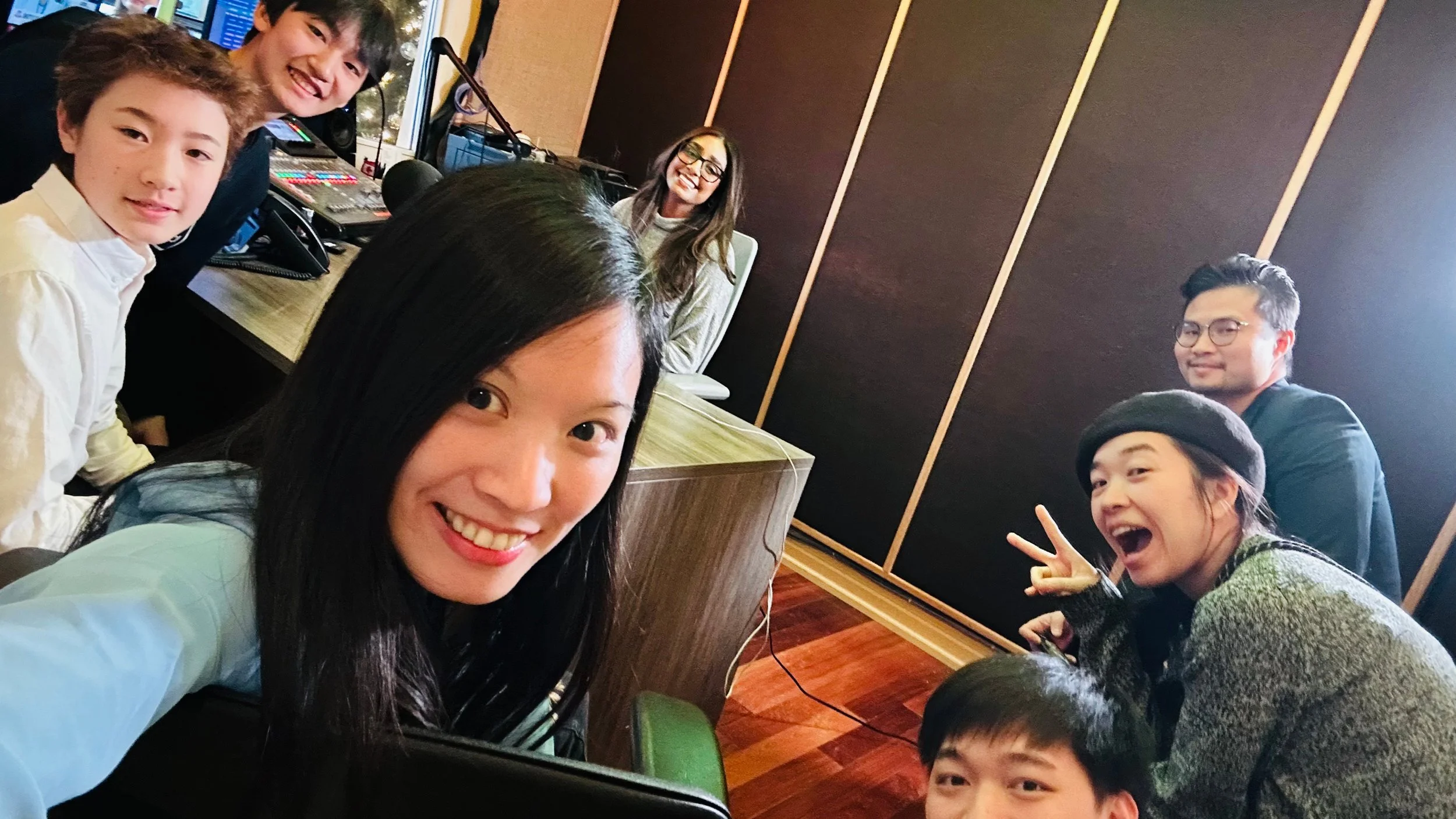 A group of young people and children smiling and making peace signs in a recording studio or radio station, with equipment and soundproofing panels visible in the background.