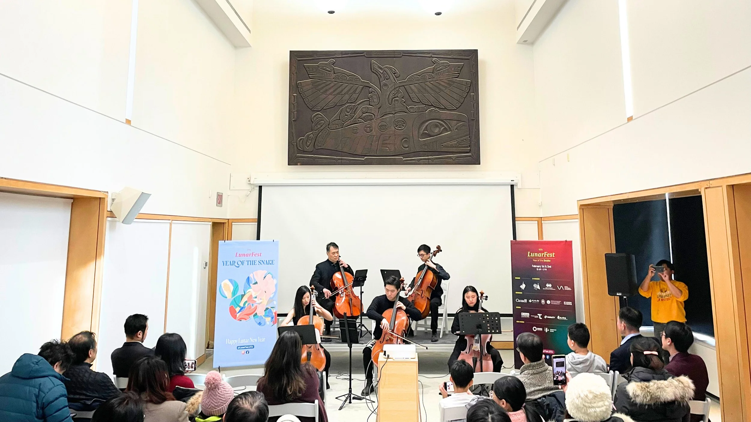 Musicians performing in a concert hall with audience seated and watching, decorated with banners and a large carved wooden artwork of a Thunderbird above the stage.