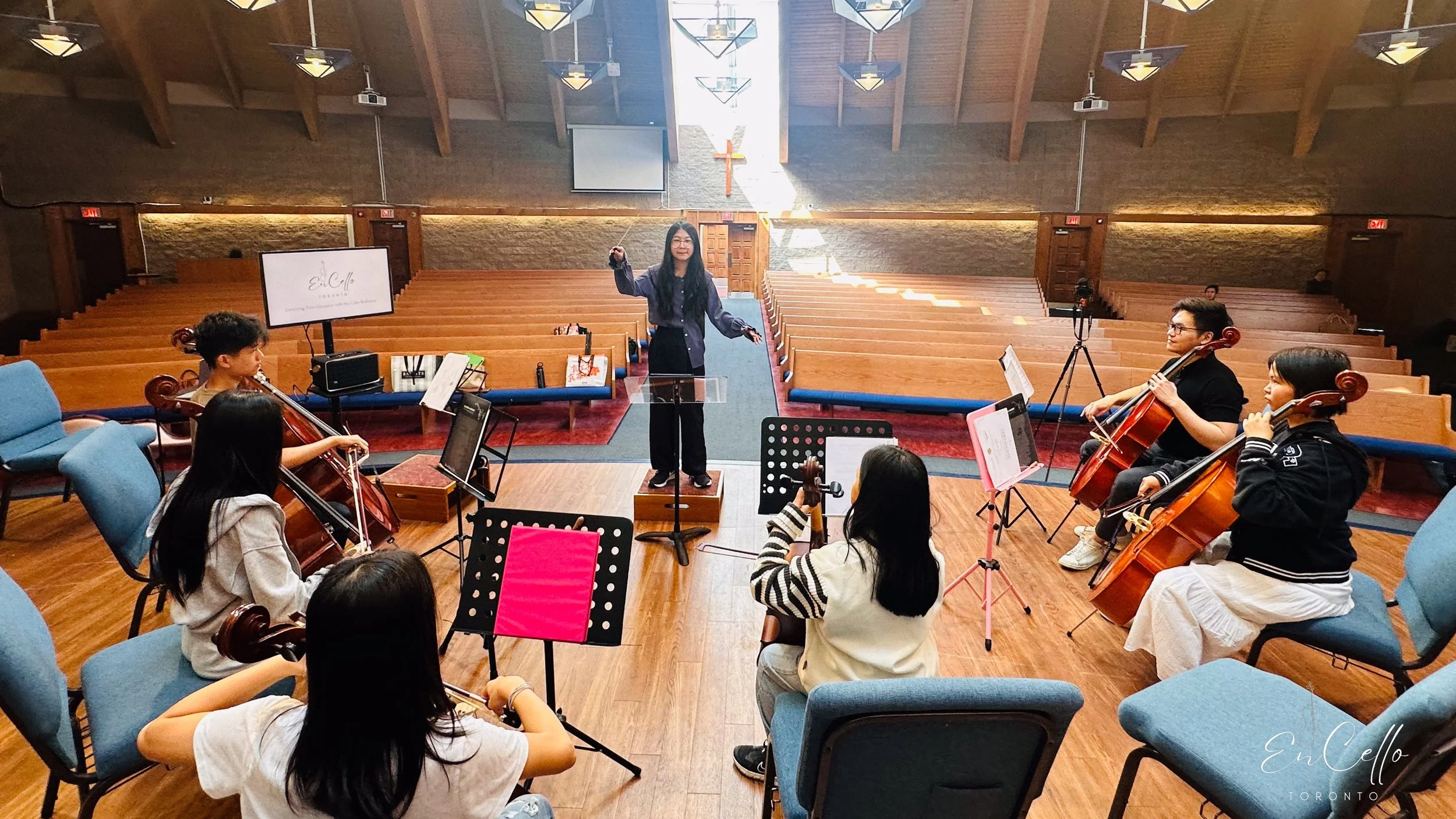 A group of musicians practicing in a church with wooden pews and a cross at the back wall. The conductor stands on a small platform, facing the musicians, with both hands raised. Several musicians are playing cellos and are seated in a semicircle aro