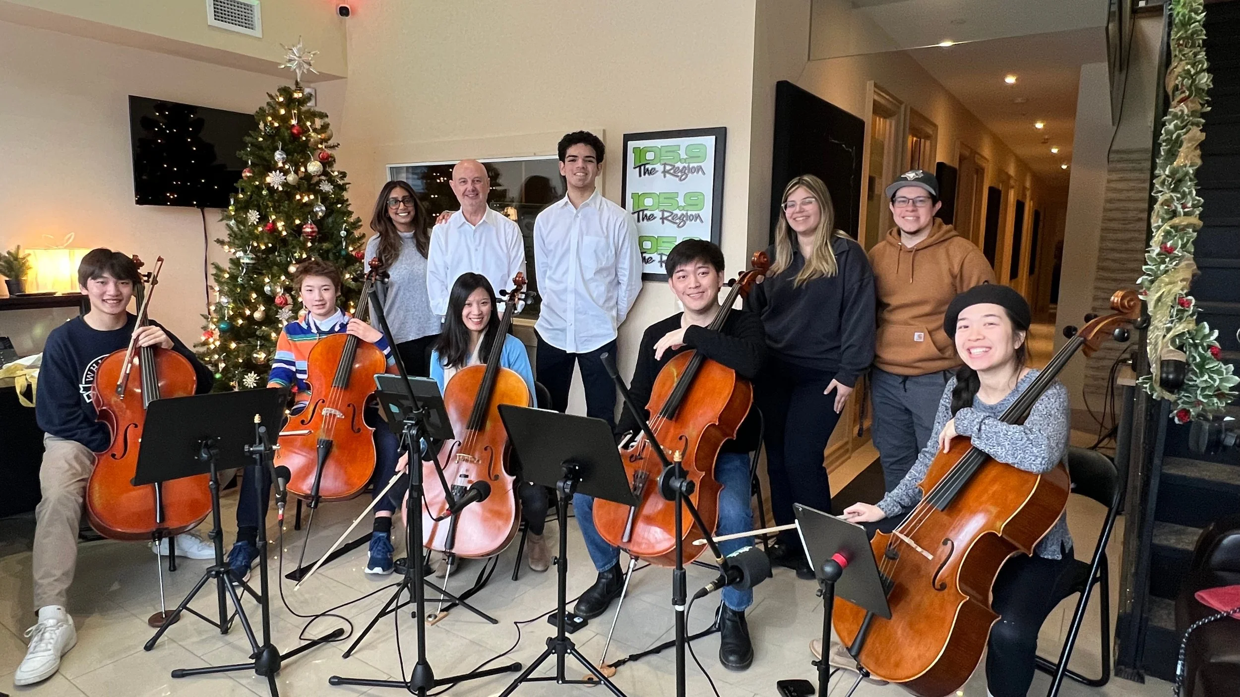 A group of young musicians with cellos and violas, smiling, standing and seated in front of a Christmas tree decorated with lights and ornaments, with a Christmas garland to the right, in an indoor room with beige walls and a television.