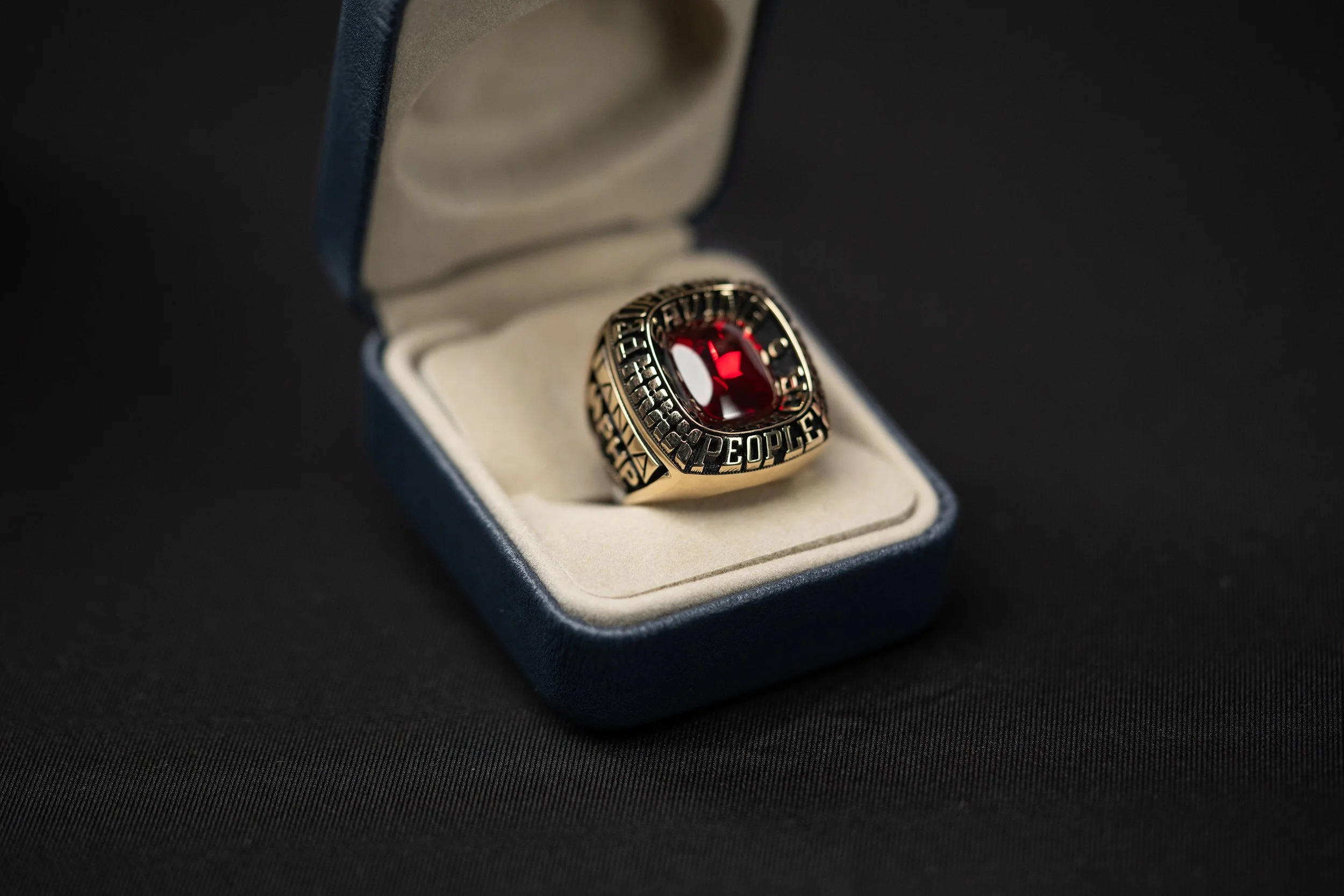 A gold signet ring with a large red gemstone in a white jewelry box with a blue exterior, set against a black background.