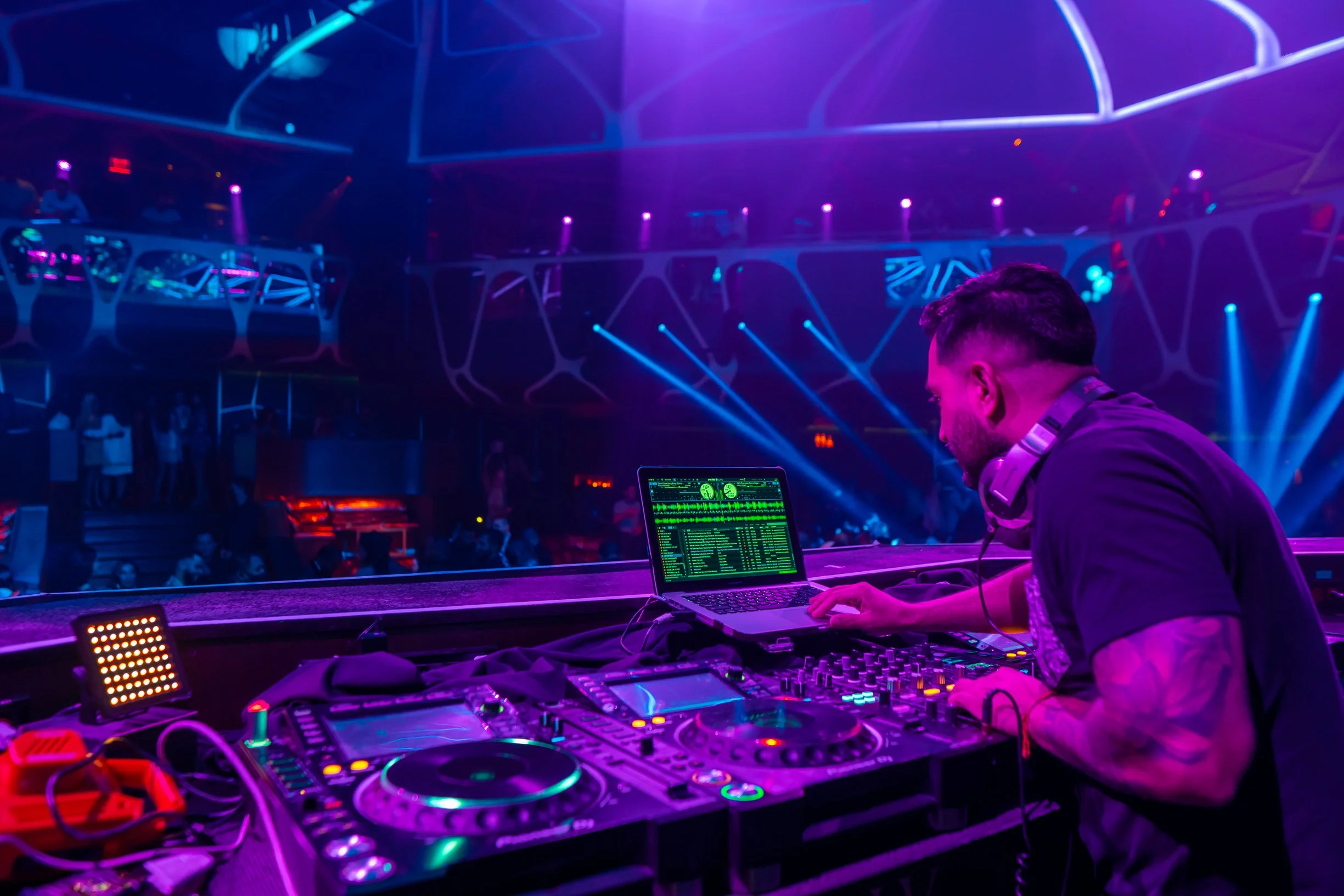 A DJ wearing headphones working on a laptop and DJ equipment in a nightclub with vibrant blue and purple lighting.