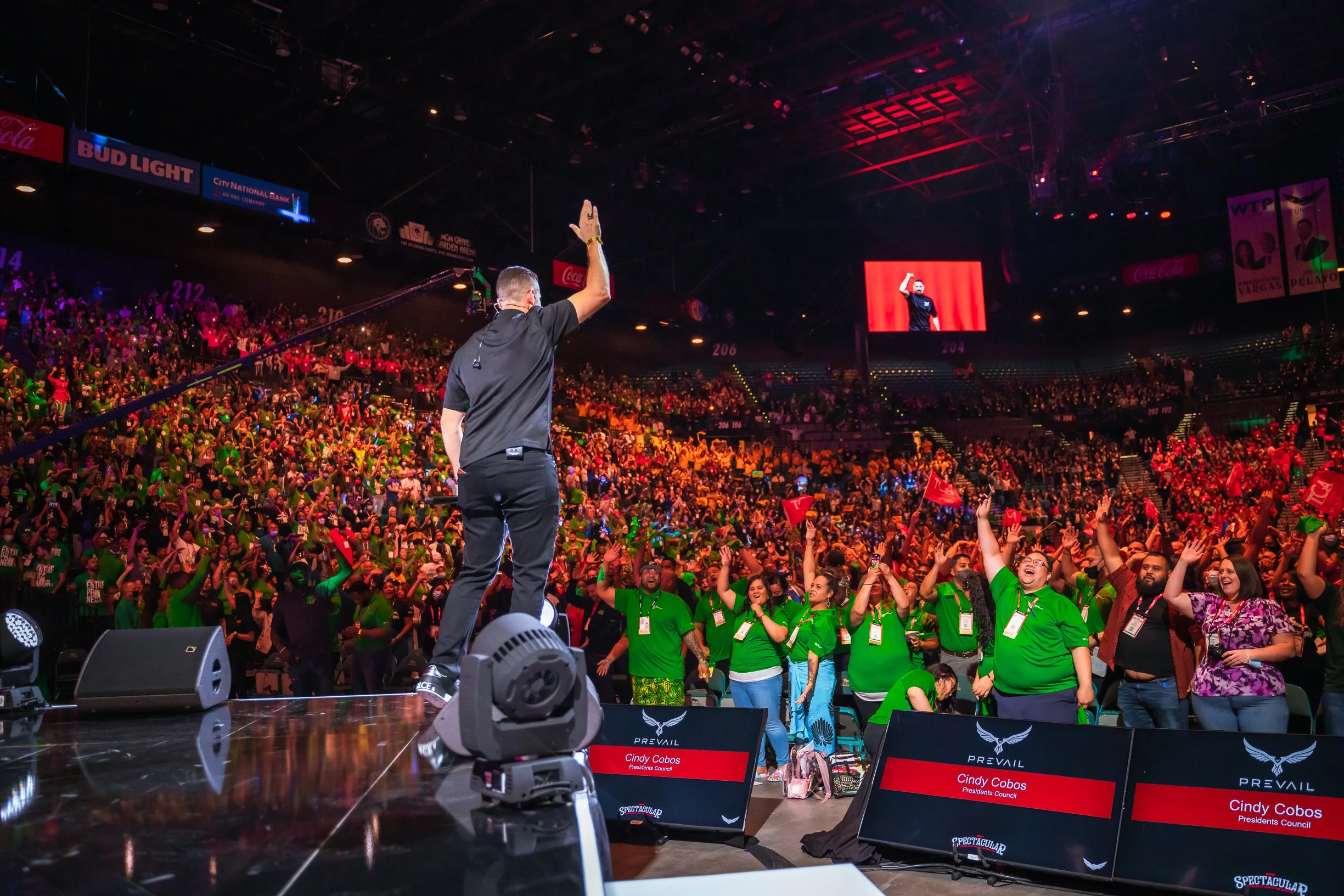 A large indoor event with a crowd audience, a man standing on stage with his hand raised, and big screens displaying his image. The audience is lively, some people have their hands raised. Signs with names and labels, including Cindy Cobos, Presidents Council, are visible in the foreground.