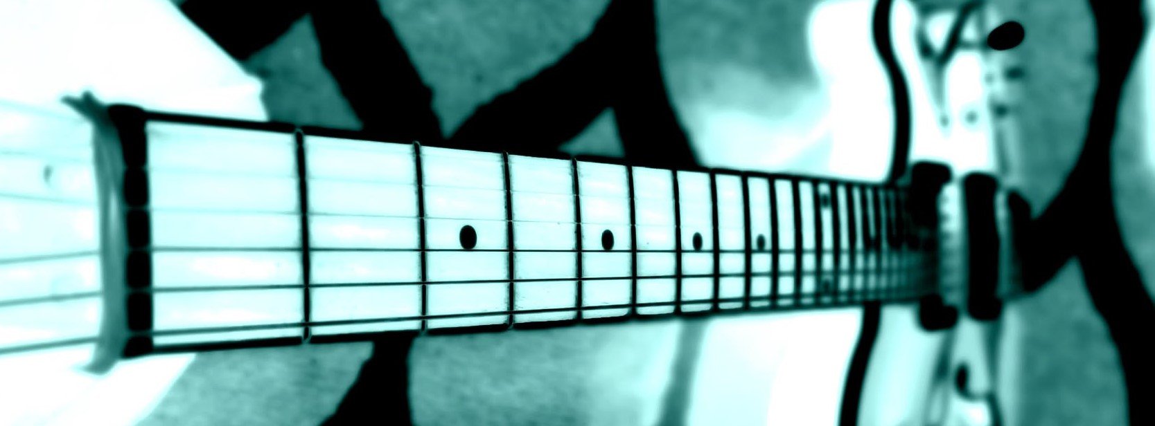 Close-up of an electric guitar neck and fretboard with black fret markers. The background is blurred, showing a guitar body and amplifier.