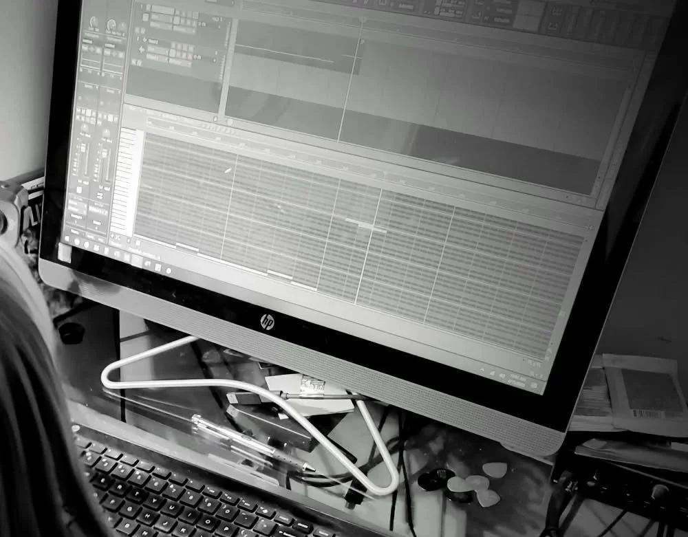 Computer monitor displaying a digital audio workstation with tracks and waveforms, surrounded by a keyboard, cables, and miscellaneous objects on a cluttered desk.