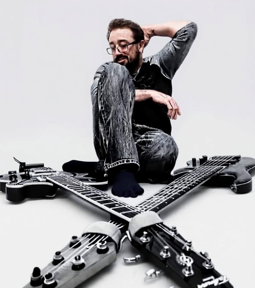 A man with glasses, a beard, and a bald spot smiling, sitting on the floor with one knee up, wearing a patterned shirt and jeans, next to a guitar-shaped coffee table.