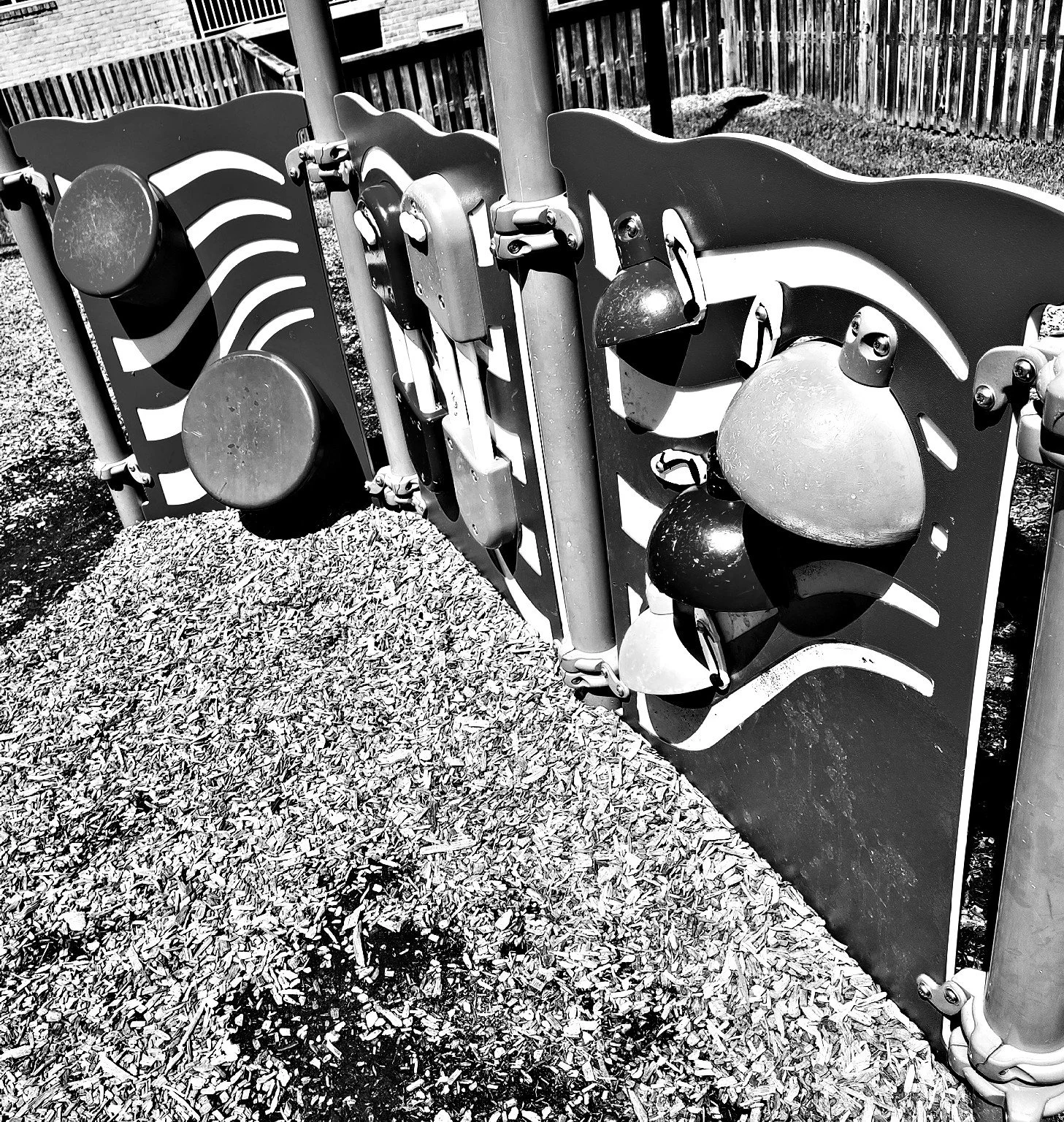 Black and white photo of a playground with climbing structures that have attached balls and curved panels, fenced in an outdoor area with wood chips on the ground.