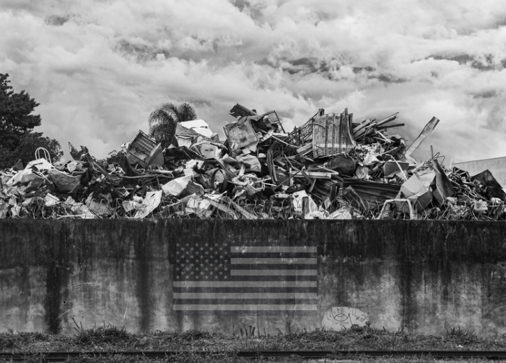 A large pile of scrap metal and debris behind a concrete wall with an American flag painted on it, under a cloudy sky.