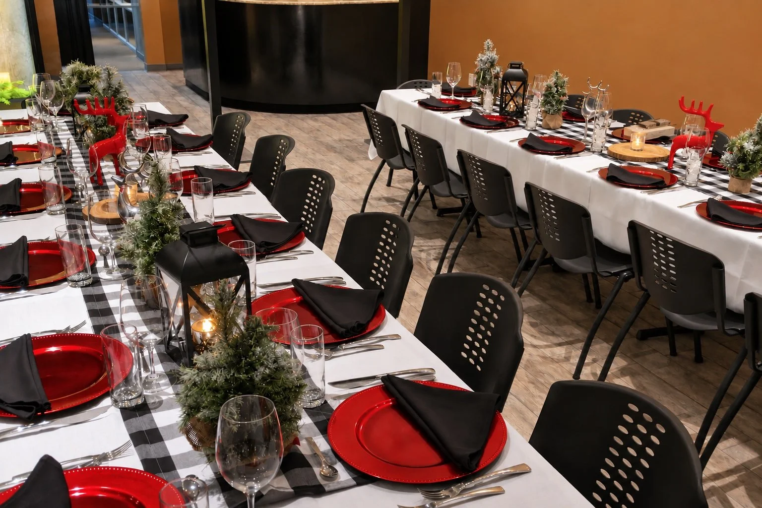 Long banquet tables set for a holiday celebration with red plates, black napkins, wine glasses, and Christmas-themed centerpieces featuring greenery and small reindeer decorations.