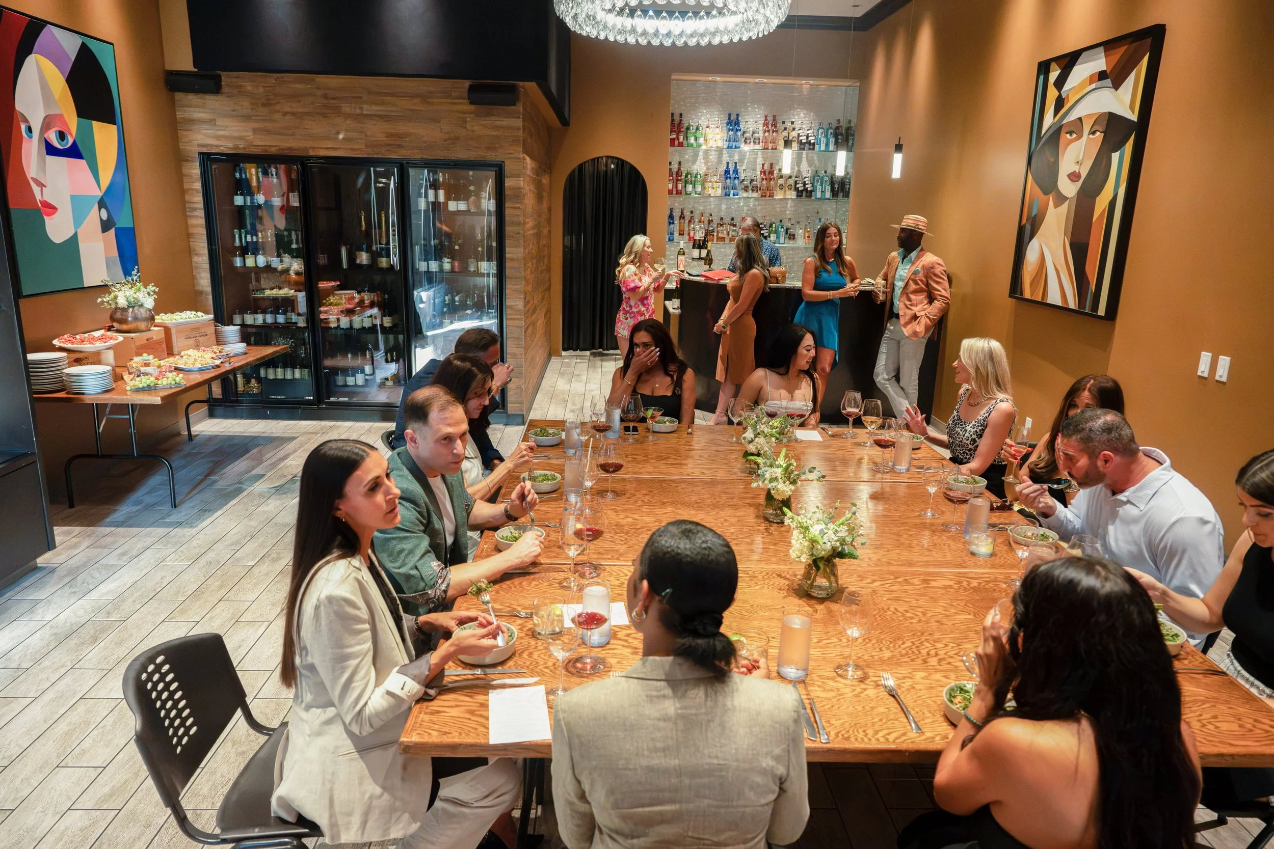 People dining and socializing in a restaurant with warm-colored walls, modern artwork, and a bar with bottles of alcohol displayed on shelves. There's a buffet of desserts and a large wooden table with people eating and talking.